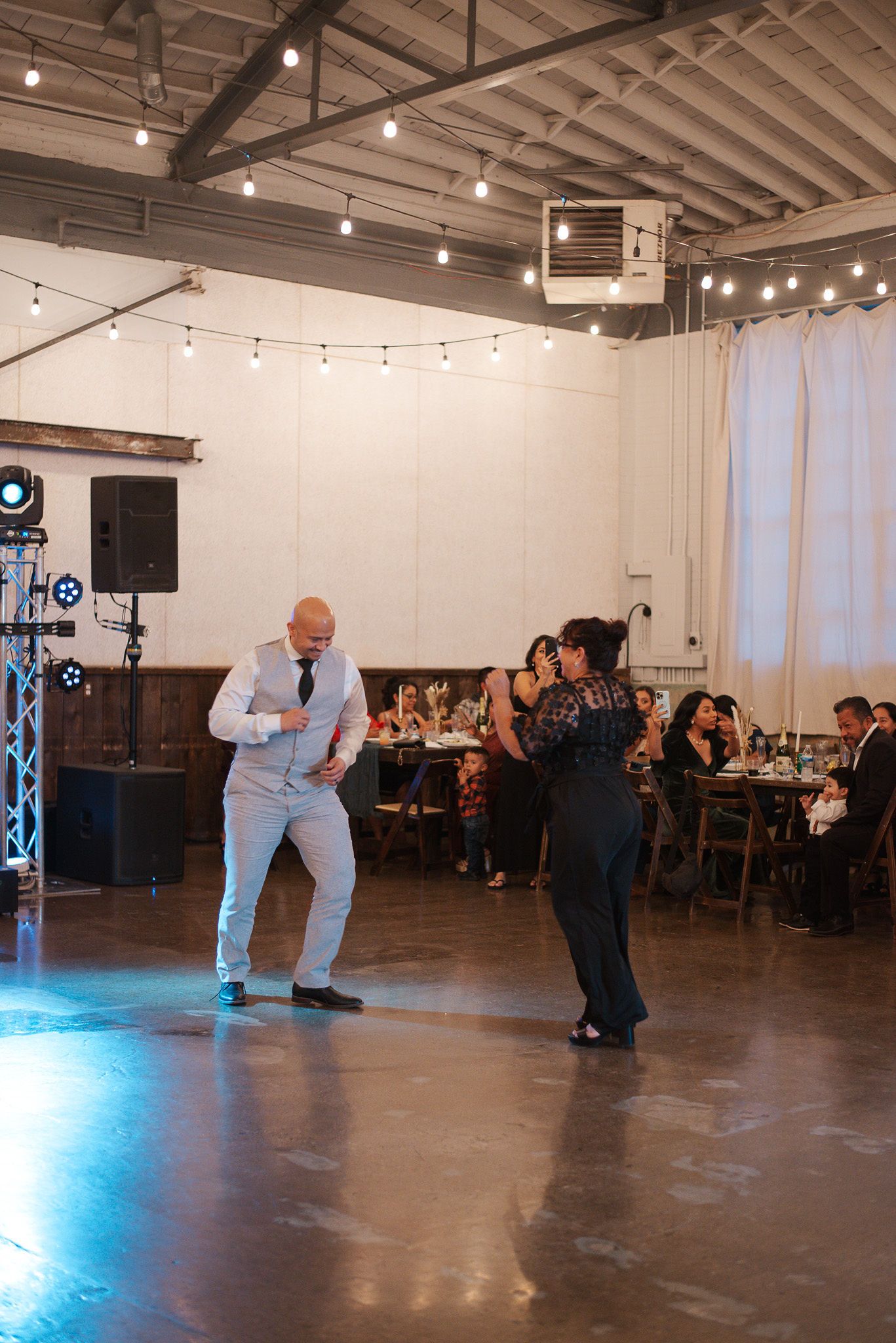 Energetic father-son dance performance at a wedding reception with string lights overhead.