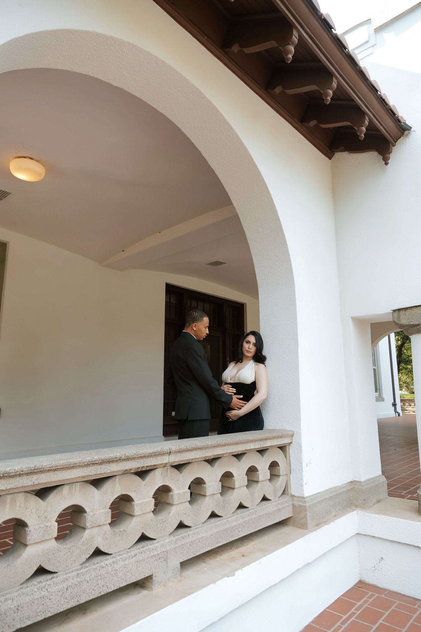 Couple in formal attire sharing a romantic moment on an elegant white arched balcony with decorative stone railing.