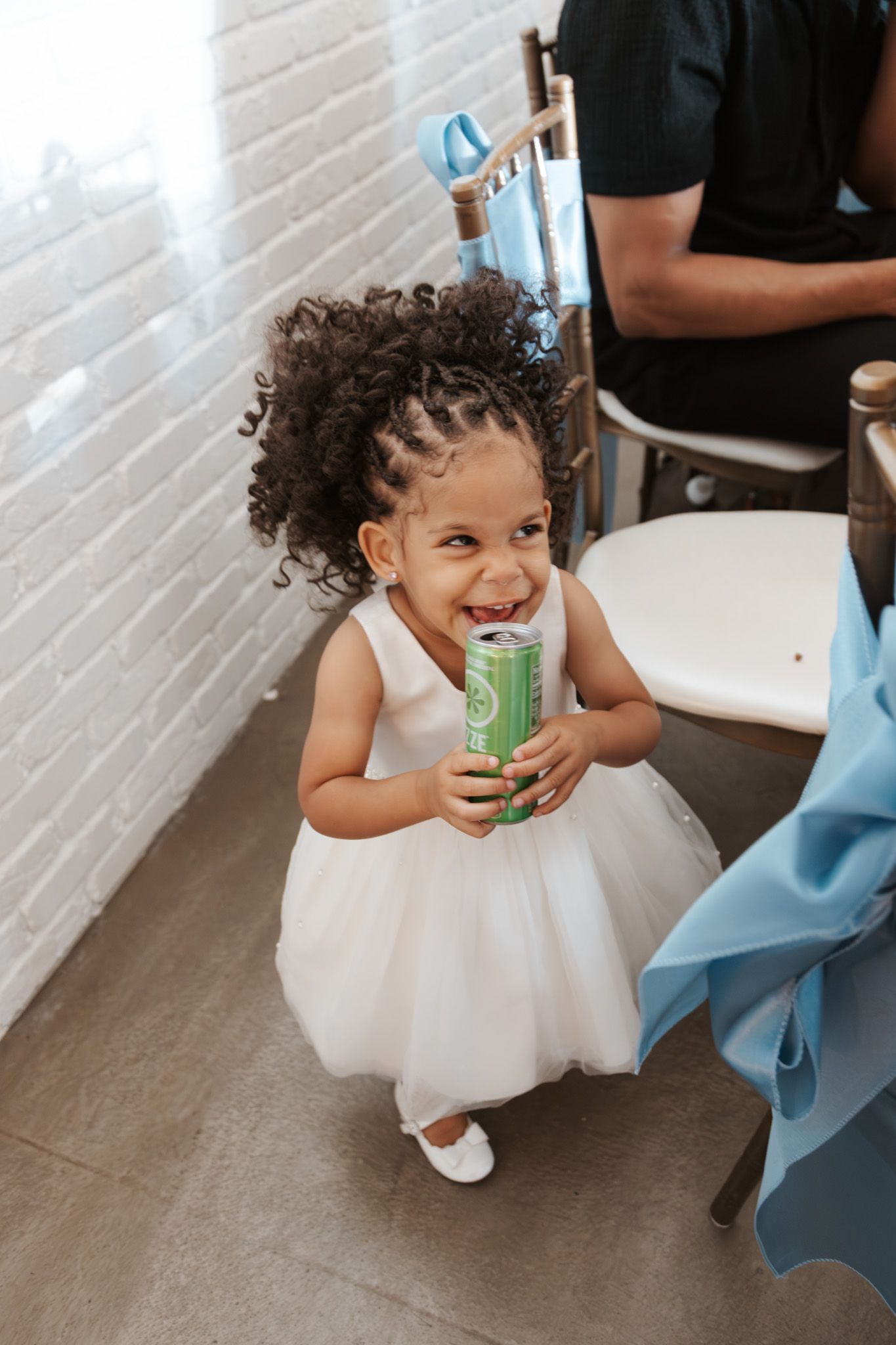 Adorable curly-haired flower girl in white dress laughs while holding a green soda can.
