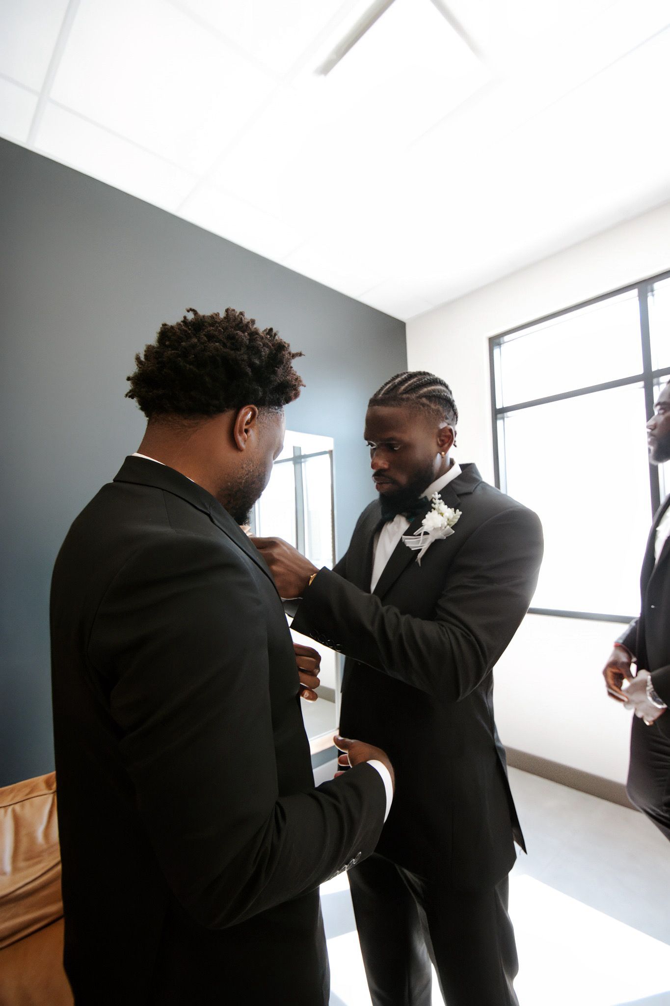 A groom adjusts his groomsman boutonniere while getting ready in a bright modern room before the wedding.