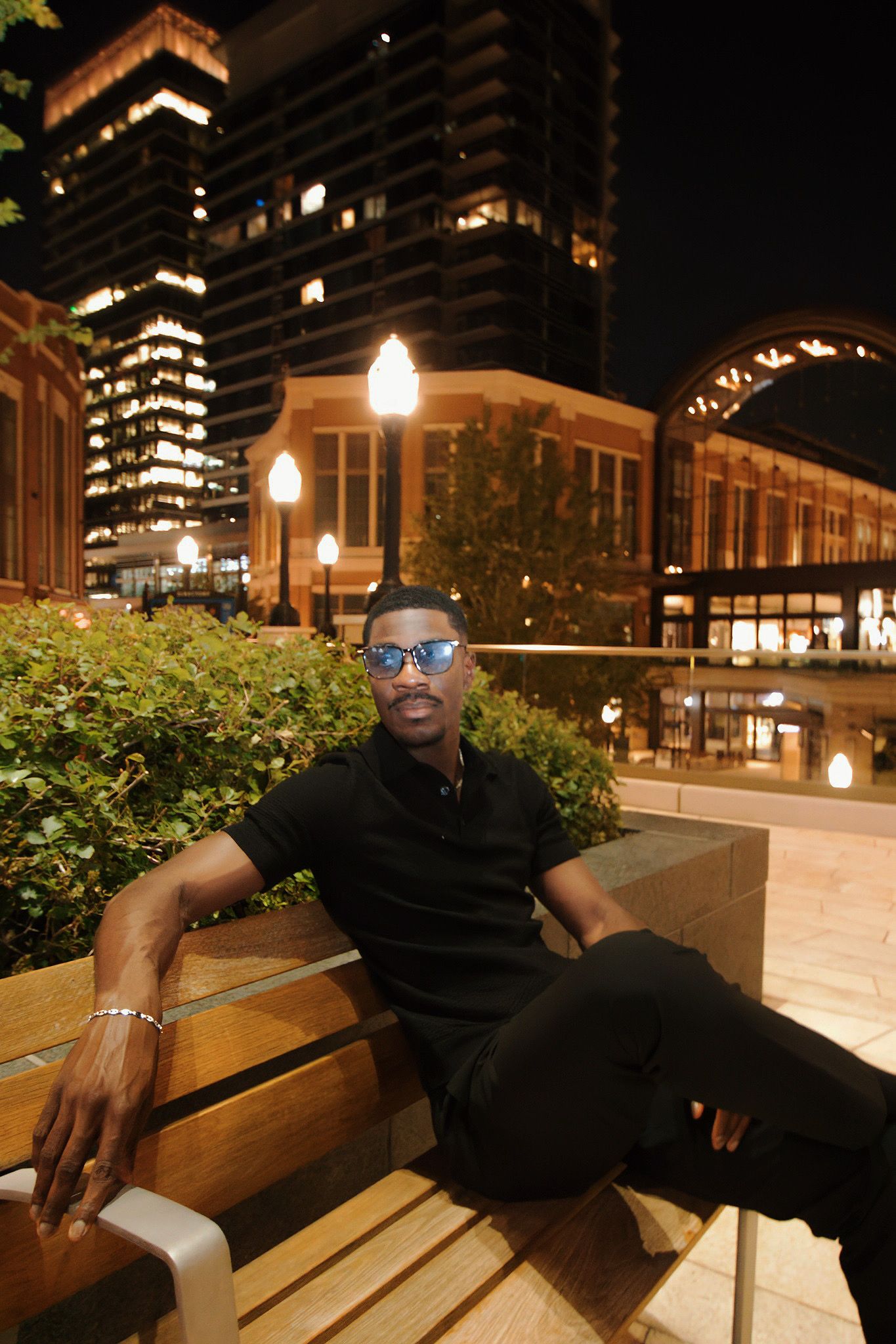 Stylish man in all-black outfit relaxing on a wooden bench in an urban city at night.
