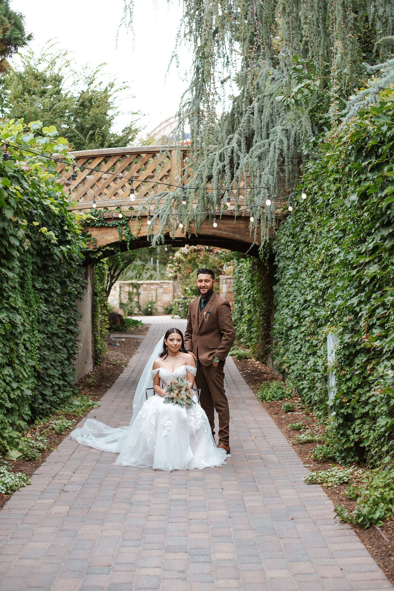 Bride and groom pose on a brick pathway surrounded by lush ivy walls and a wooden bridge at an outdoor wedding venue.