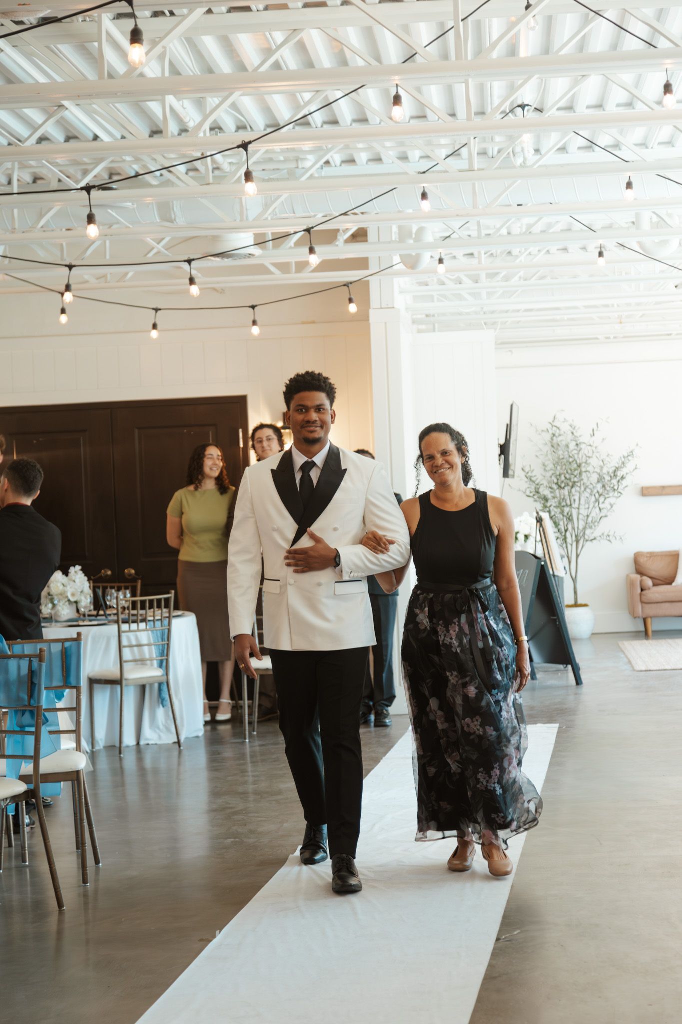 Groom in white tuxedo jacket walks down aisle with woman in floral dress at elegant indoor wedding venue.