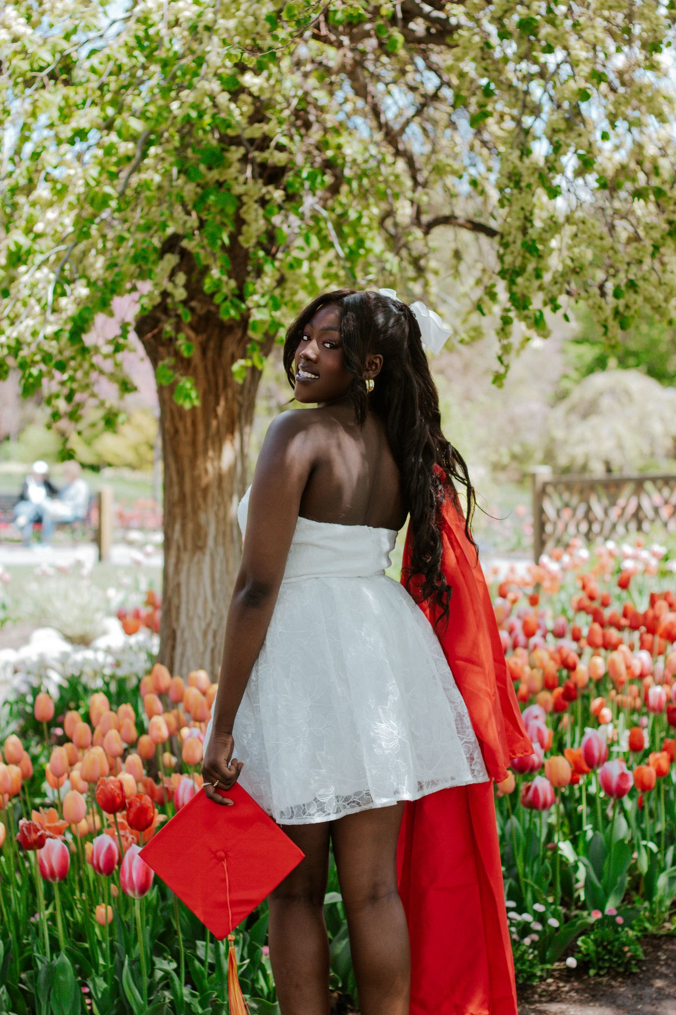 Smiling Black woman in white dress holding red graduation cap among colorful tulips in bloom.