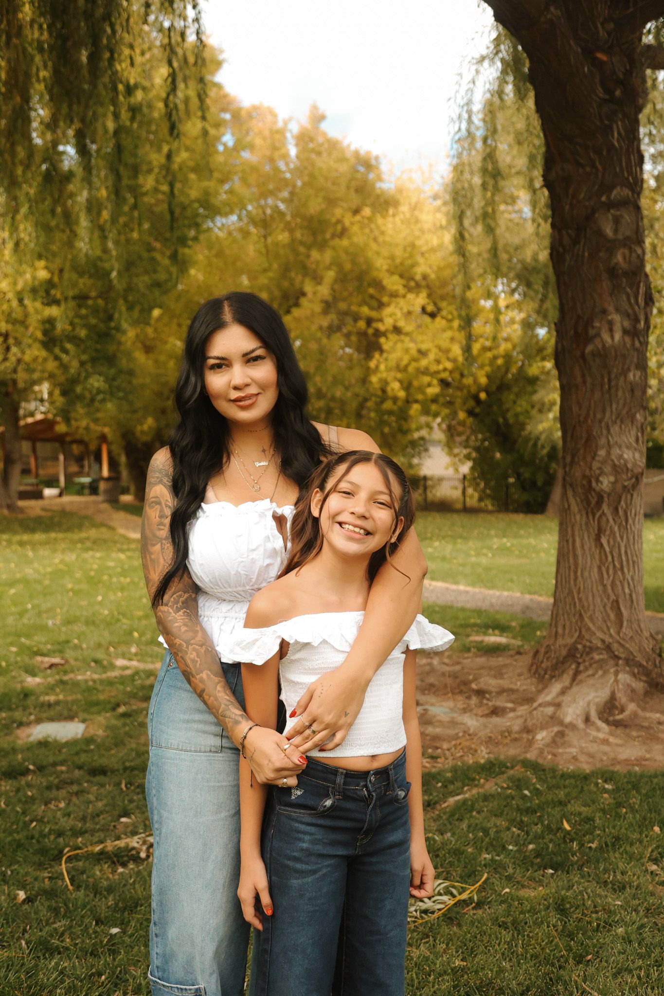 Mother and daughter smiling together in a sunny autumn park under a willow tree.