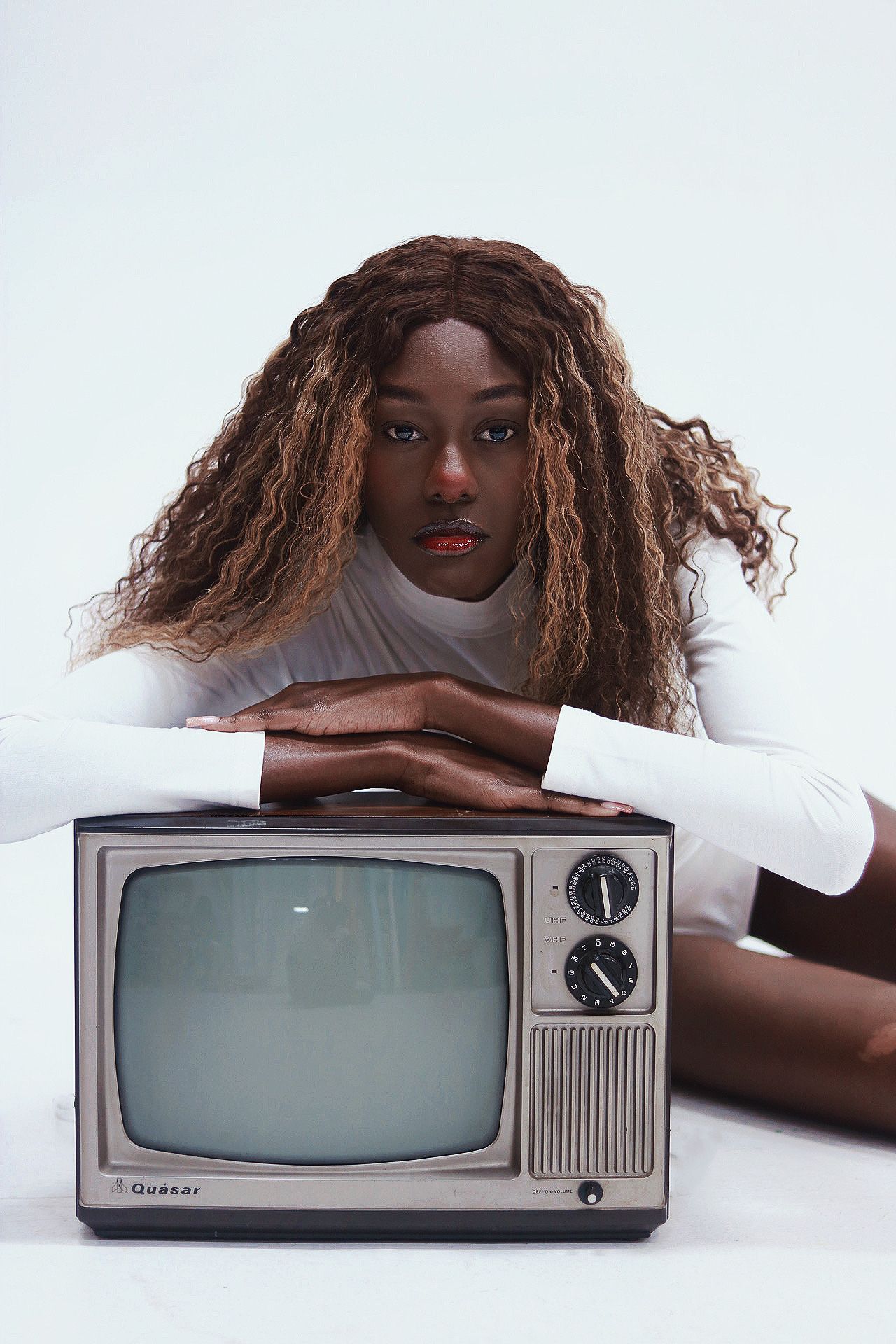 Black woman with long curly highlighted hair leaning on vintage Quasar TV against white background.