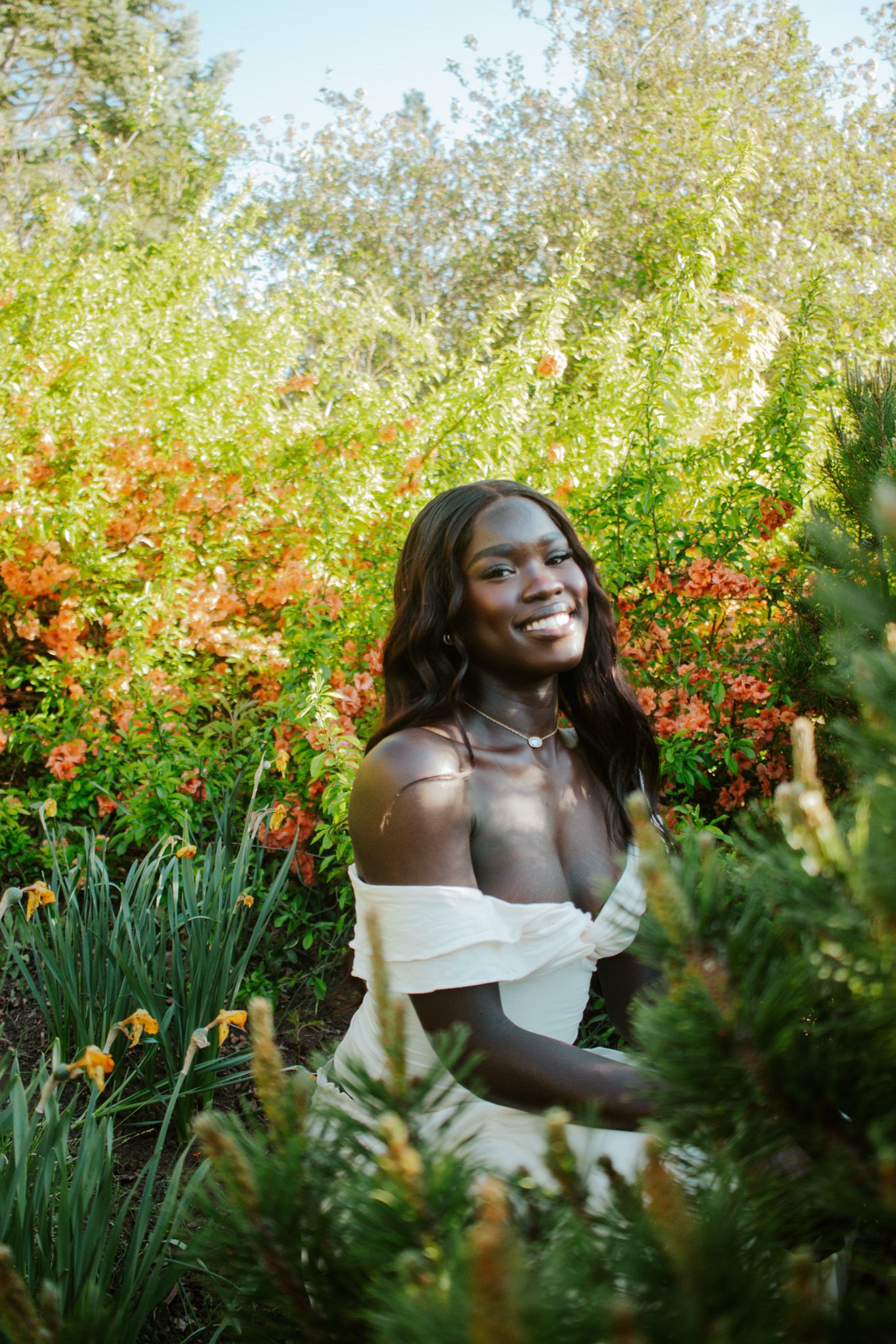 Smiling woman in white off-shoulder dress sitting among vibrant orange and red garden flowers.