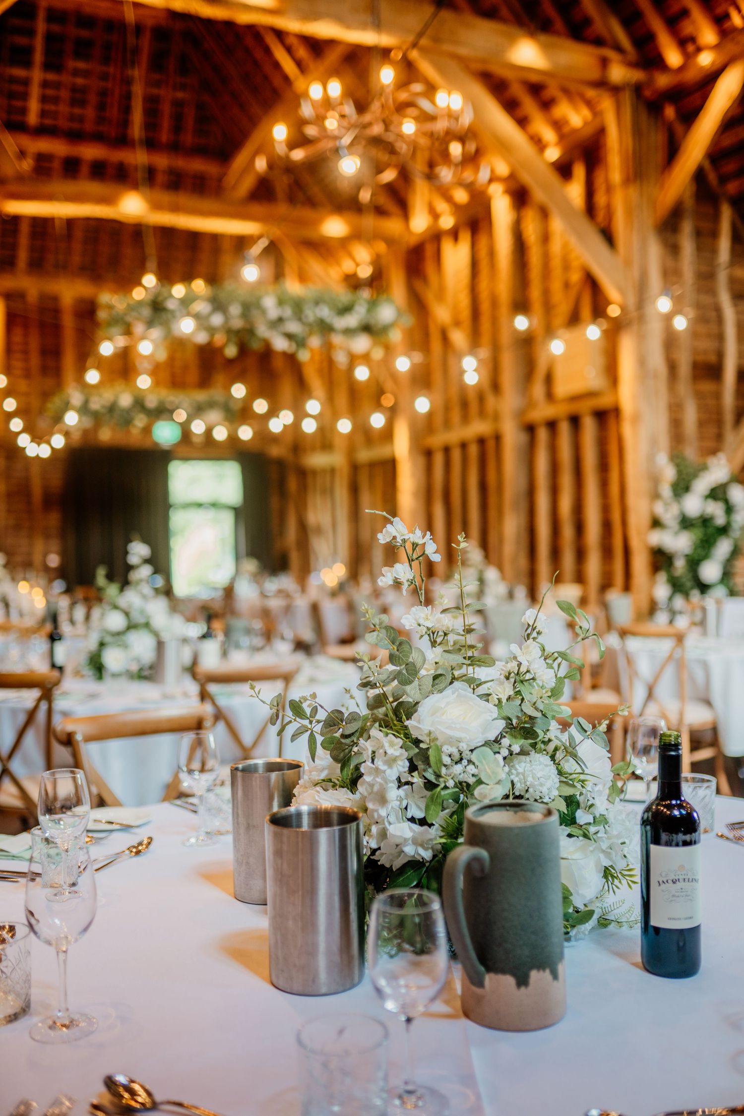 Rustic barn wedding reception setup with white linens, floral centerpieces and string lights creating warm ambiance.