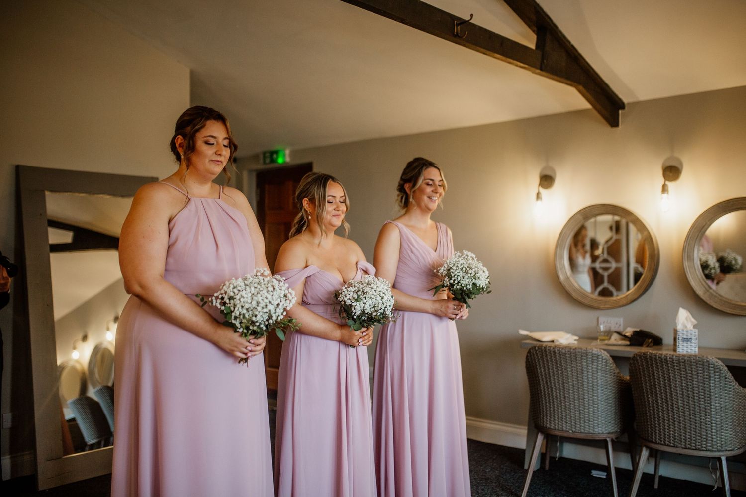 Wedding party in pink dresses preparing in an elegant venue with mirrors and wooden beams.