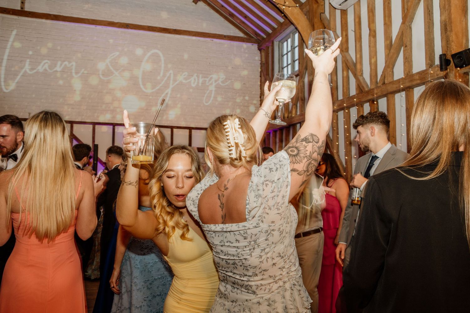 Wedding guests dancing and celebrating on a dance floor inside a rustic barn venue with exposed wooden beams.