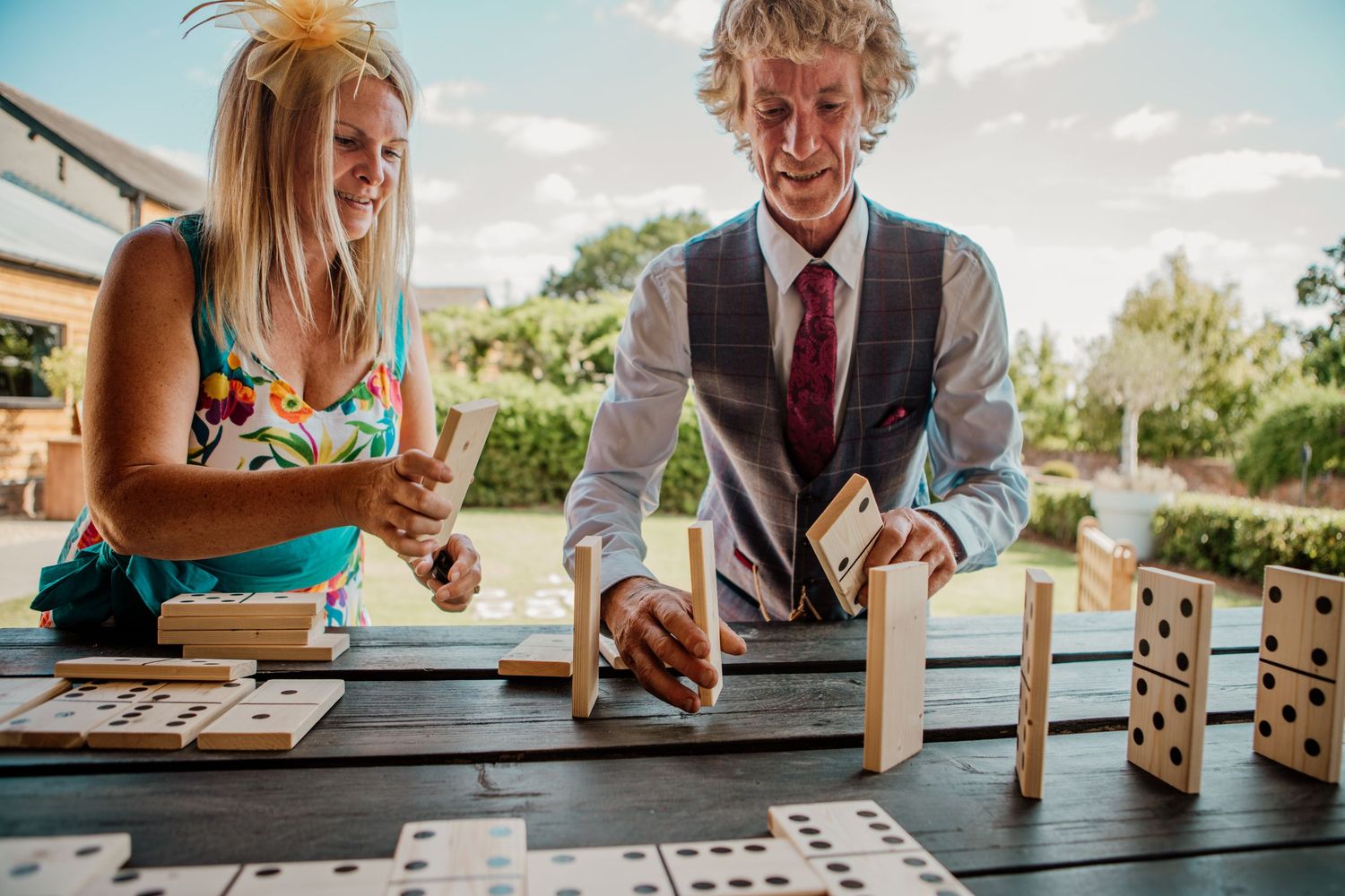 People playing a board game outdoors on a sunny day, with multiple players gathered around a wooden table.