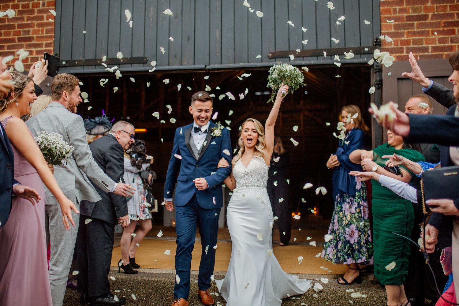 Newlyweds exit their ceremony while guests shower them with white confetti petals against a brick backdrop.