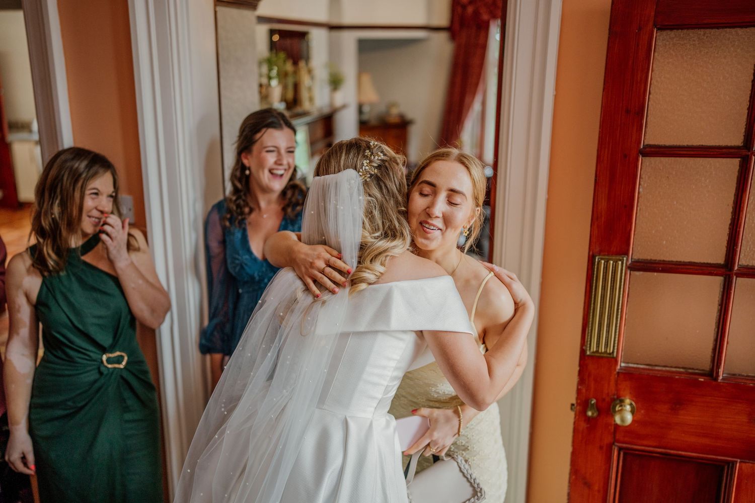Wedding party celebrates joyfully in a sunlit room with bridesmaids in emerald green dresses.