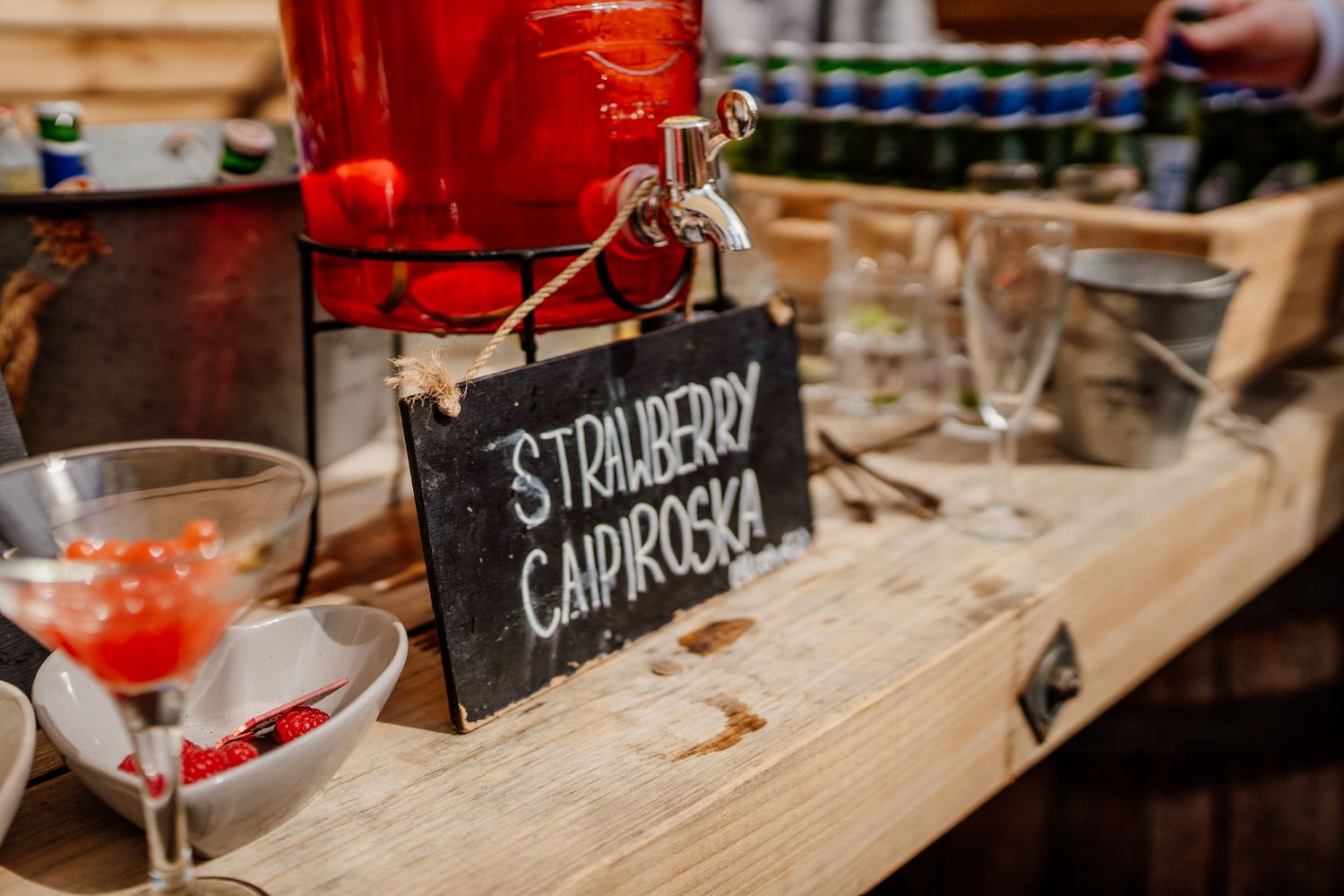 A rustic bar counter with cocktail glasses and handwritten drink menu signs displaying Strawberry and Caipiroska options.