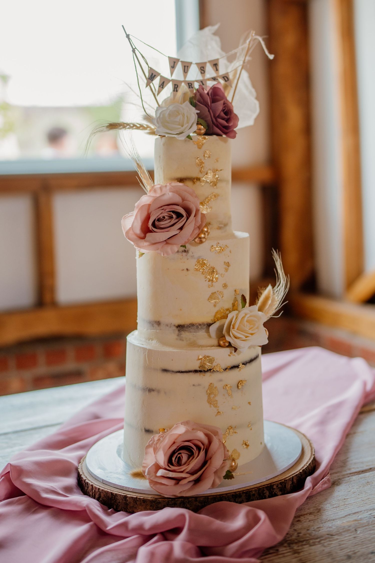 Elegant white wedding cake with dusty pink roses and gold accents displayed on wooden table with pink linen.