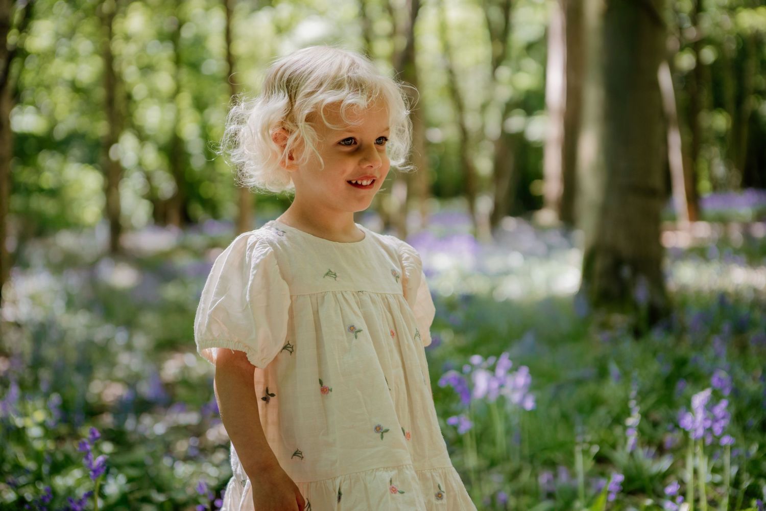 Child in white dress explores blooming bluebell woodland during spring season.