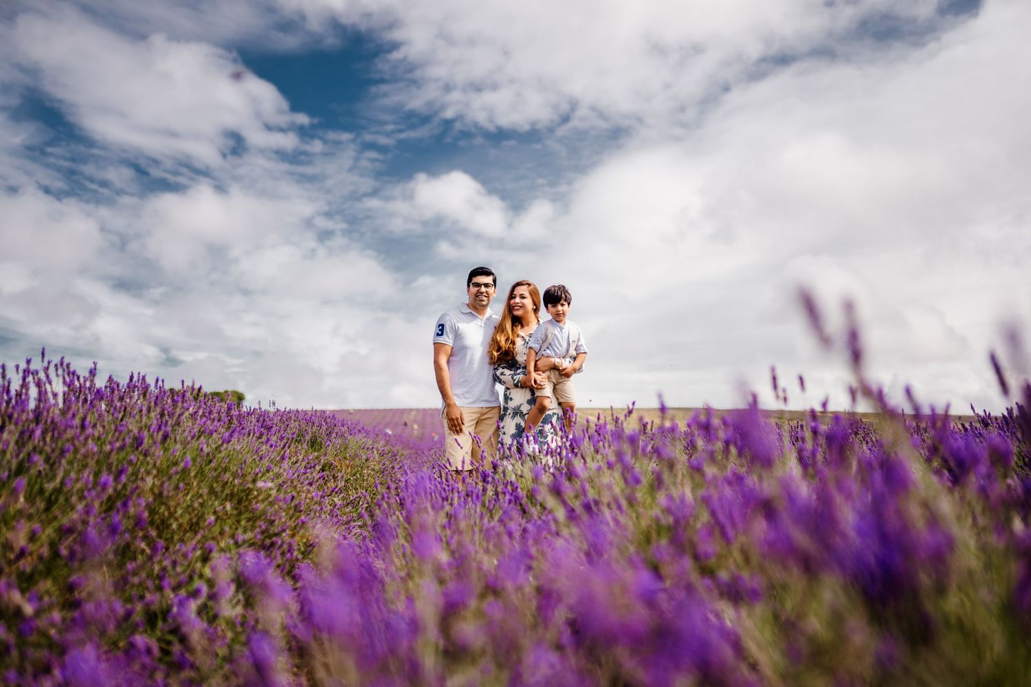 Family of three standing in a vibrant purple lavender field under a partly cloudy blue sky.