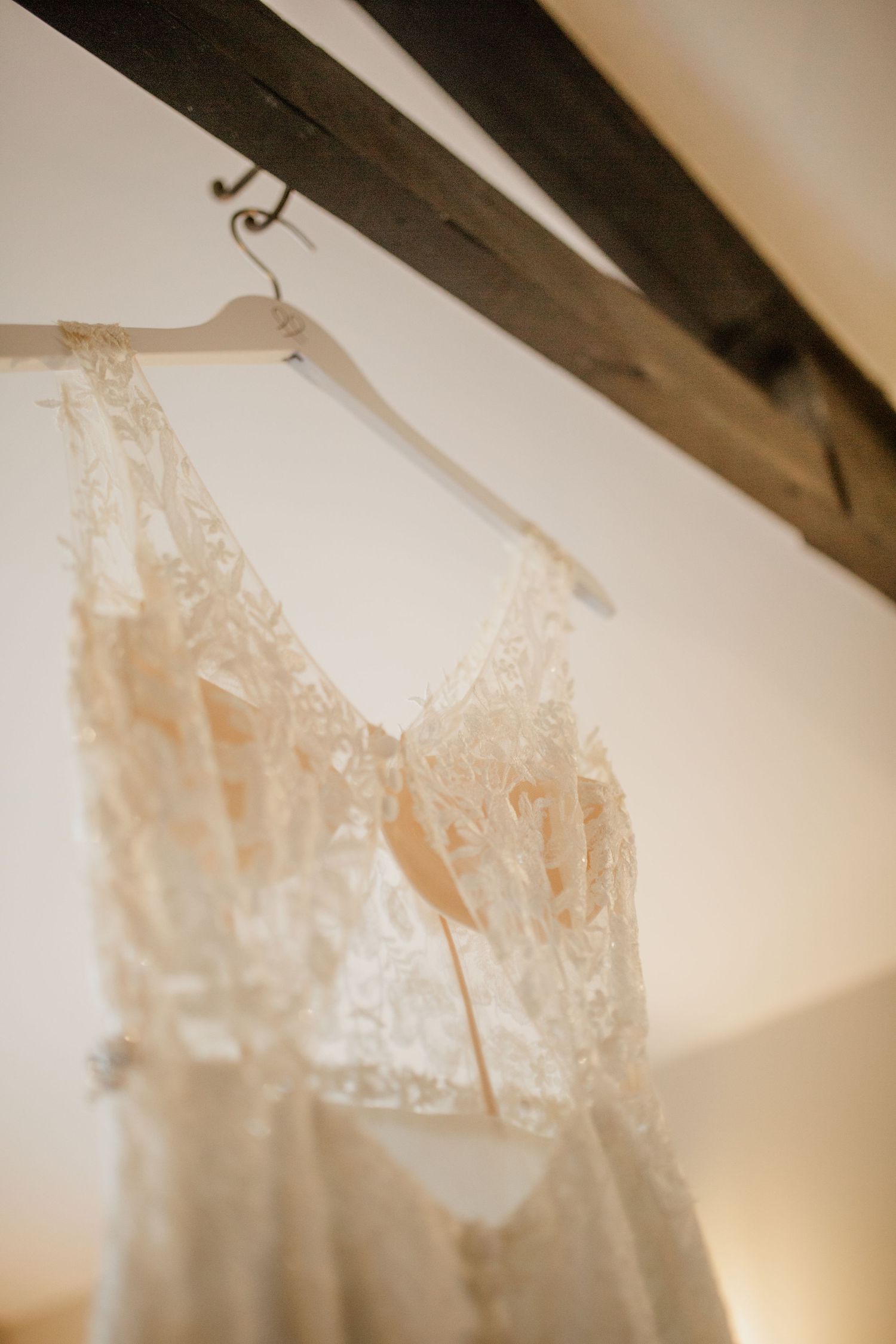 Delicate lace wedding dress hanging against a white wall with exposed wooden beams.