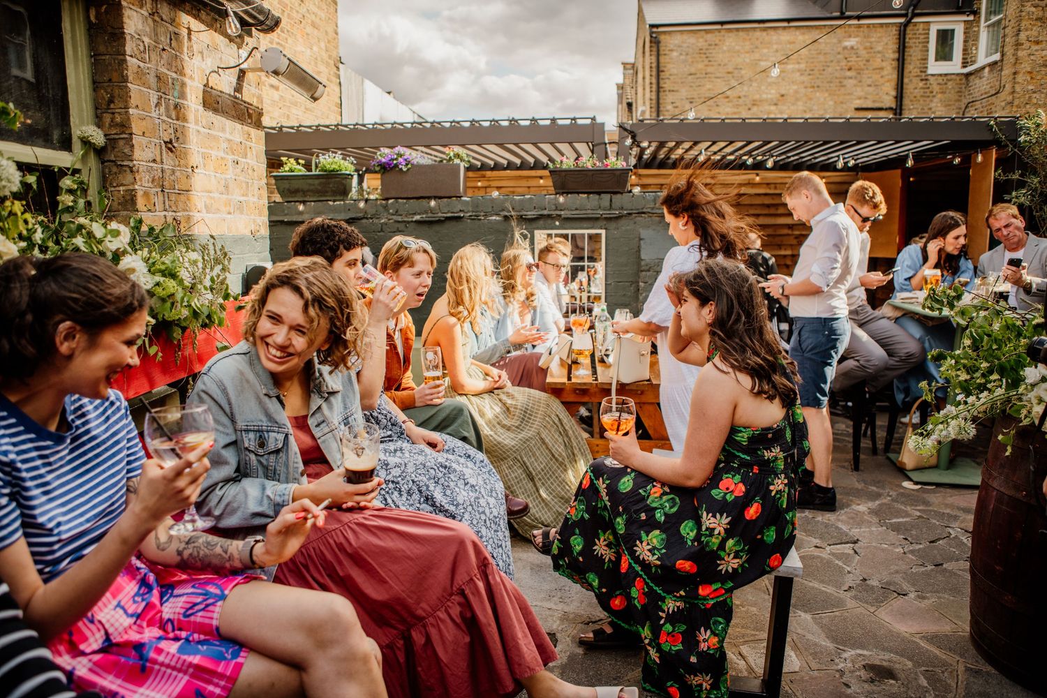 A group of friends enjoying drinks and conversation at an outdoor patio with stone walls and floral decorations.