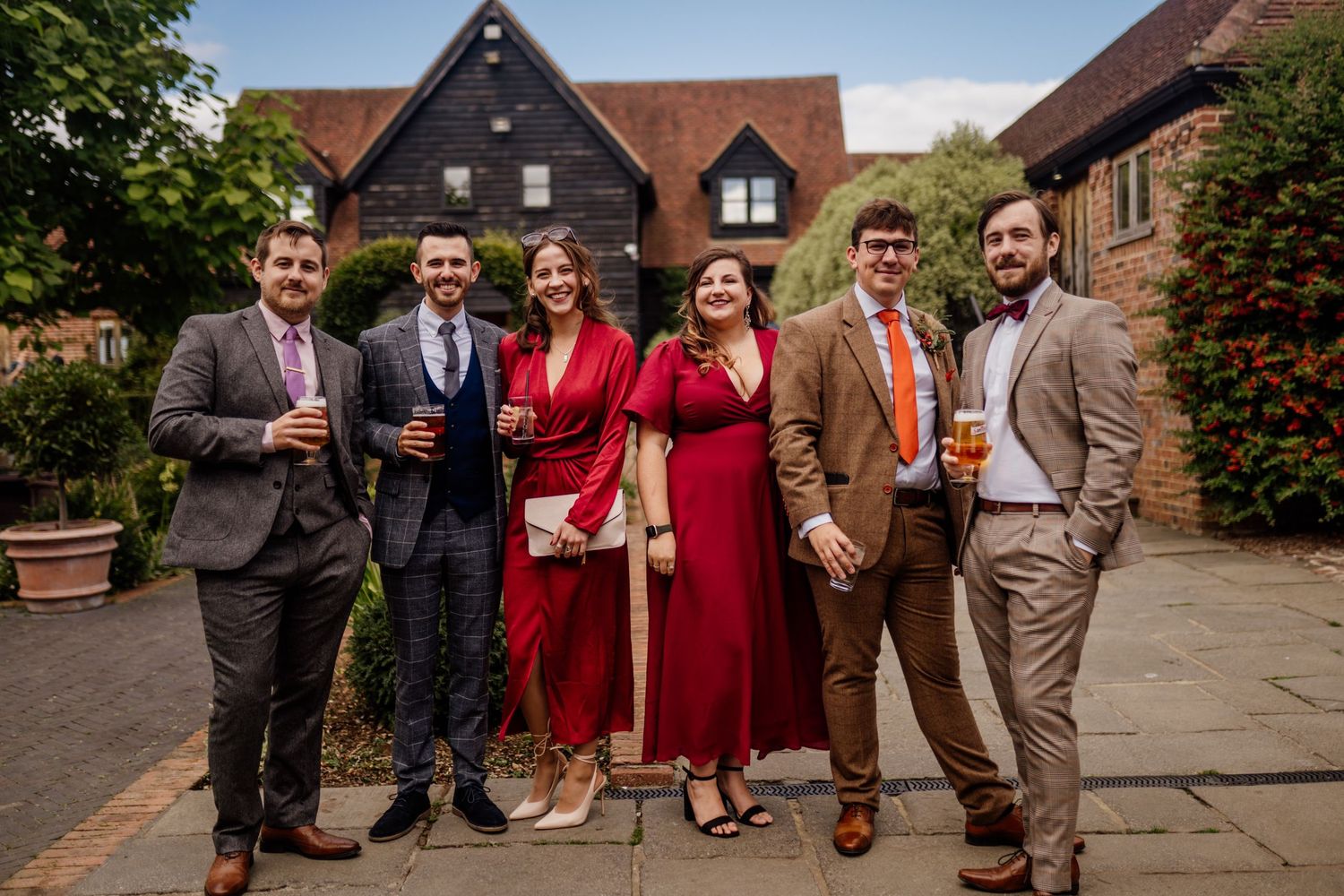 A group of wedding guests in formal attire gather outside a rustic brick building on a sunny day.