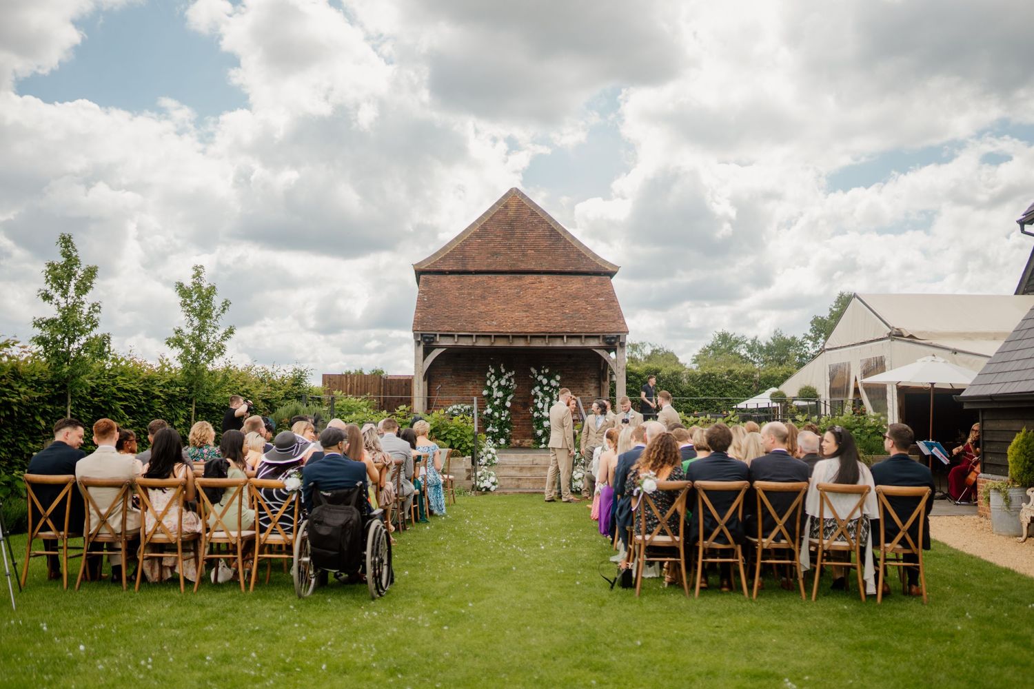 Outdoor wedding ceremony setup with wooden chairs on a green lawn under a rustic gazebo and cloudy summer sky.