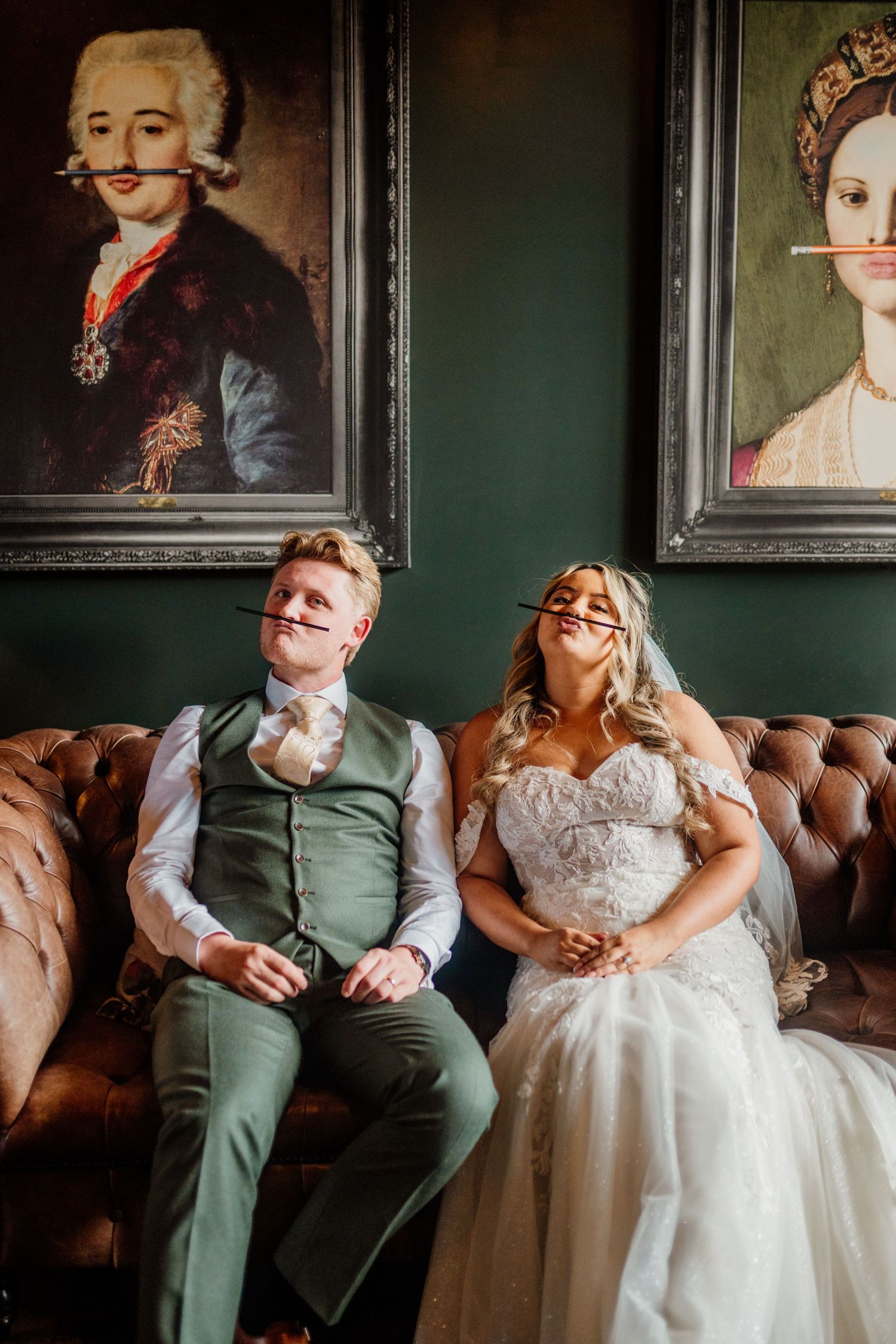 A couple in wedding attire relaxes on a vintage brown leather couch with ornate portrait frames on the wall behind them.