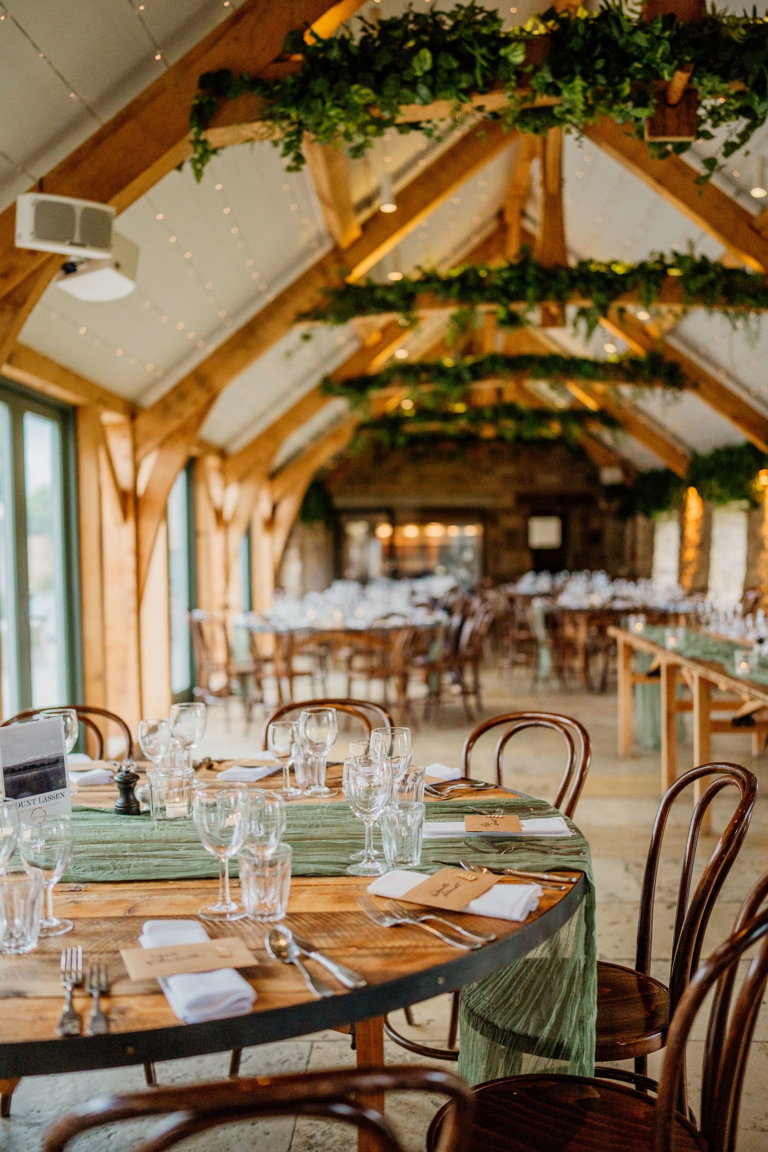 Rustic wedding reception venue with wooden beams, hanging greenery, and elegant table settings in natural light.