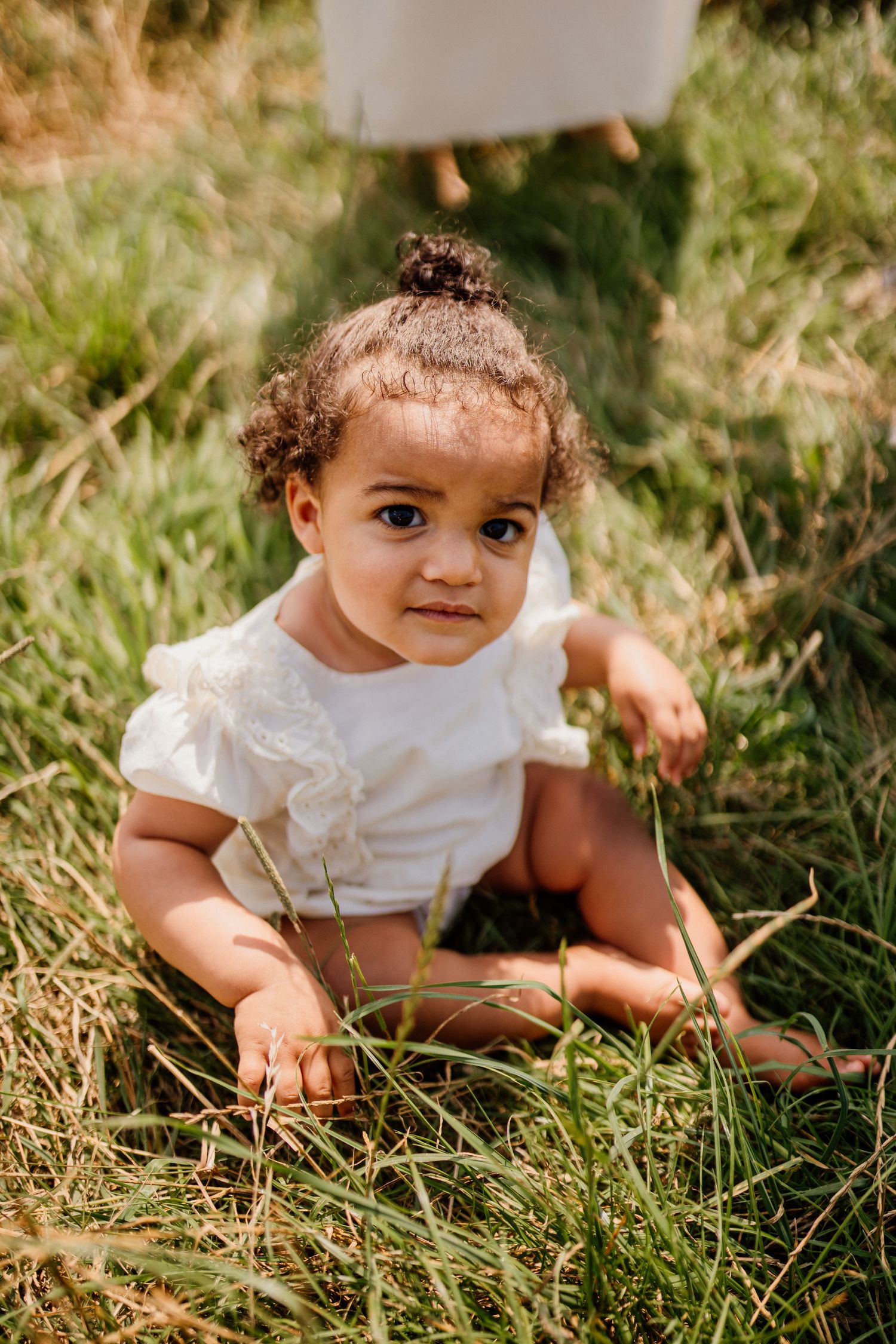 A small child in a white dress sits among tall grass in natural lighting.