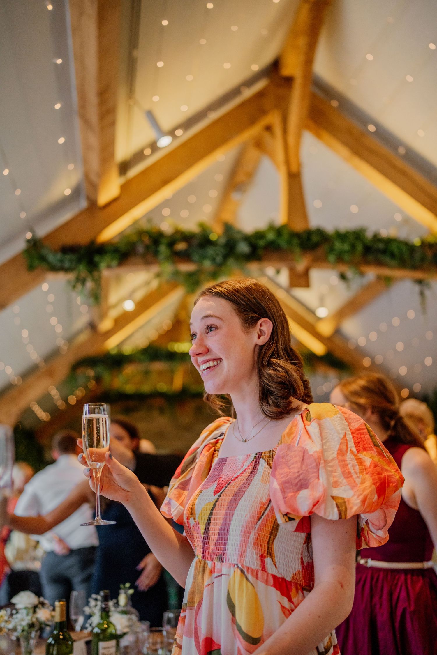 Guest in coral dress raises champagne glass during wedding reception under twinkling lights.