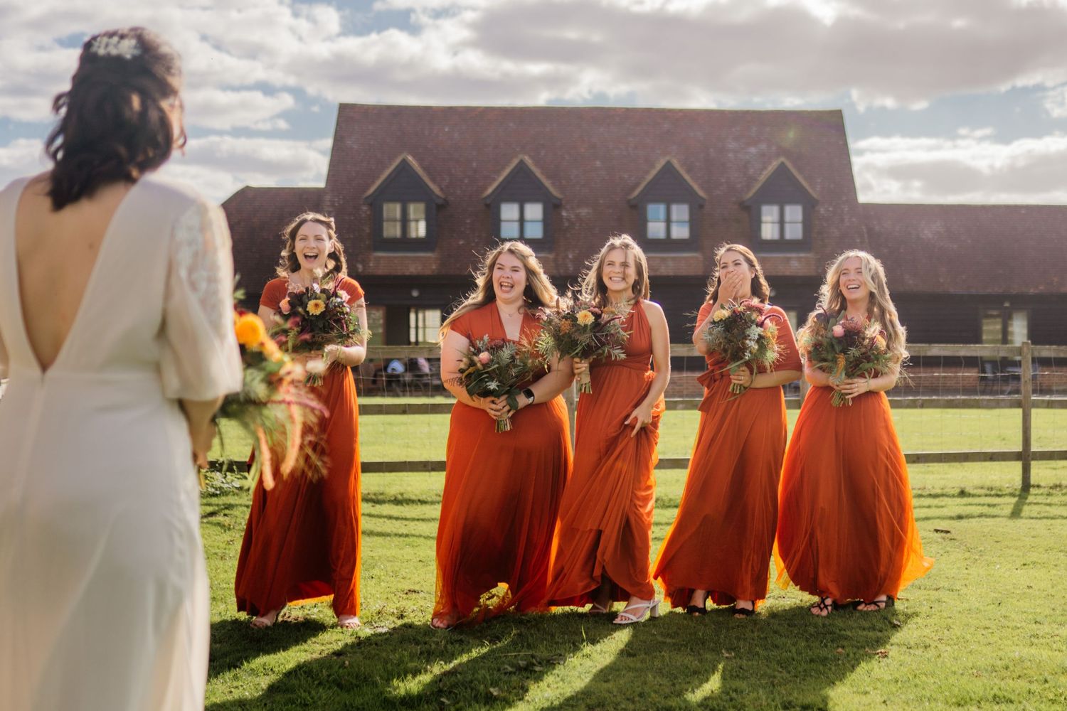 Bridesmaids in bright orange dresses stand in a line on a grassy lawn outside a rustic venue during sunset.