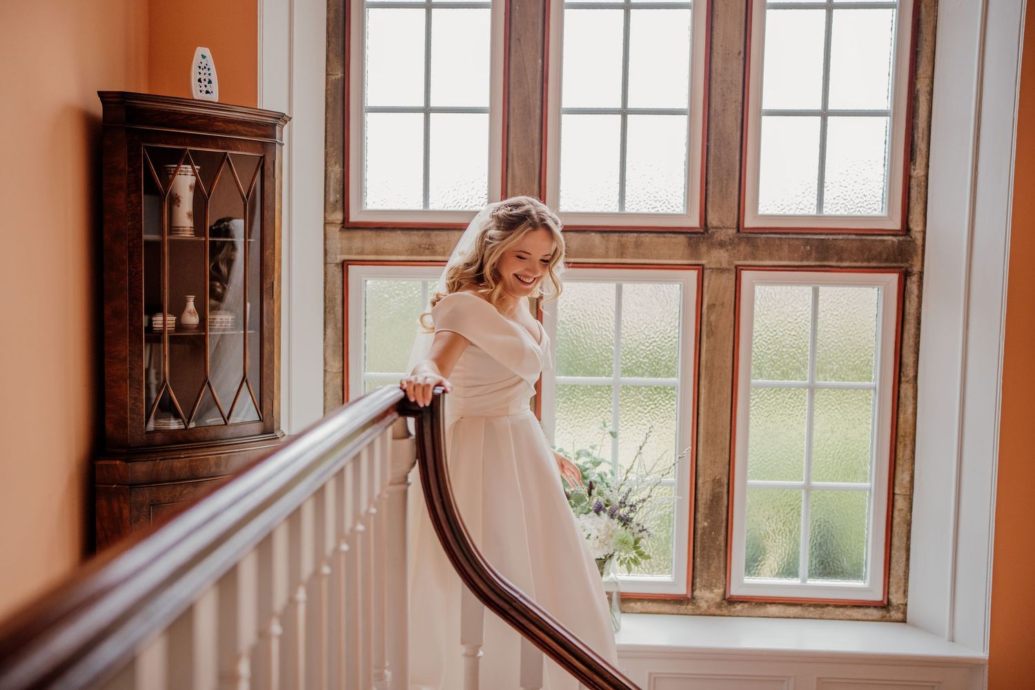 Elegant bride in white dress stands by window in rustic room with natural light streaming through wooden frames.