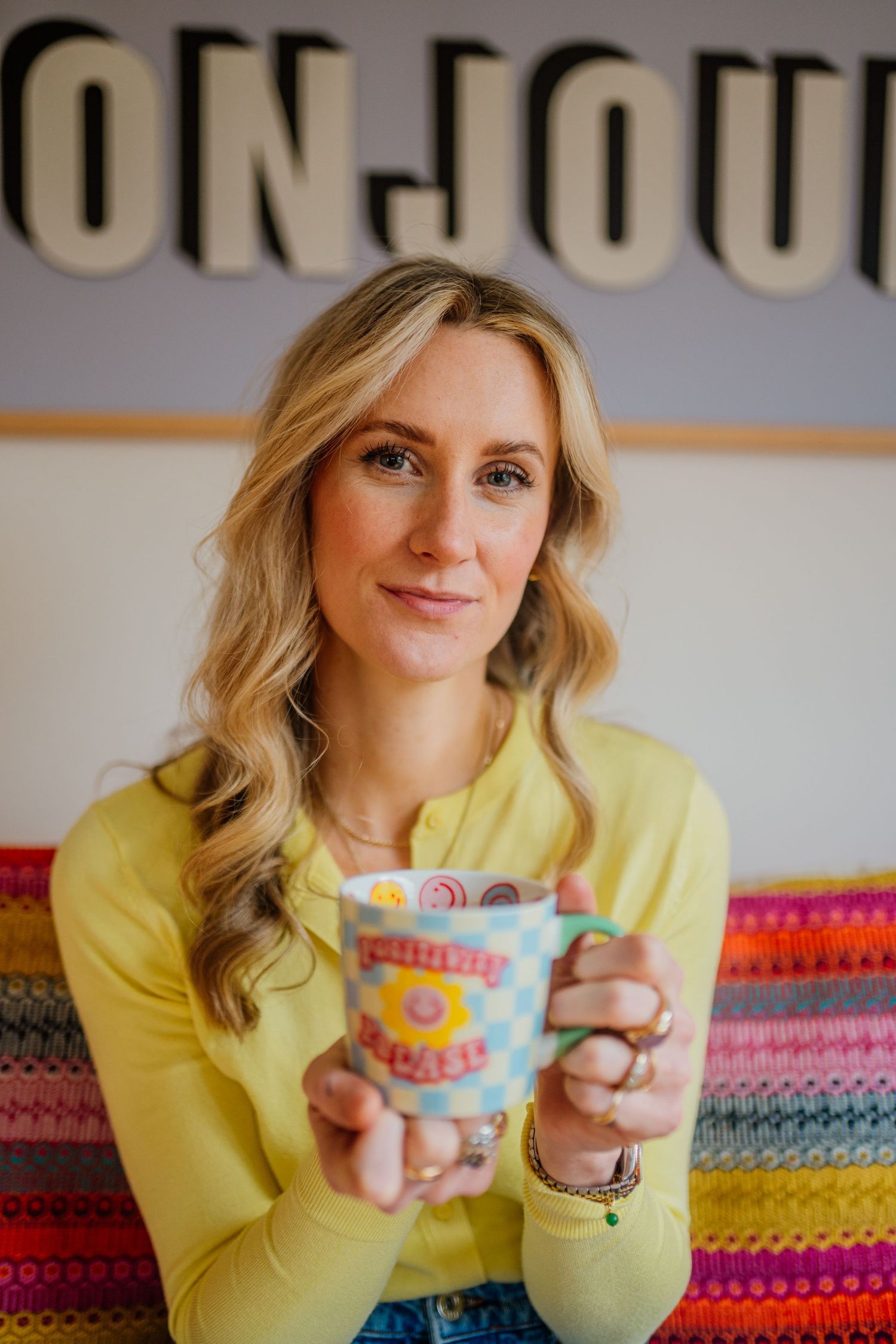 Person in yellow sweater holding colorful ceramic mugs against striped background.