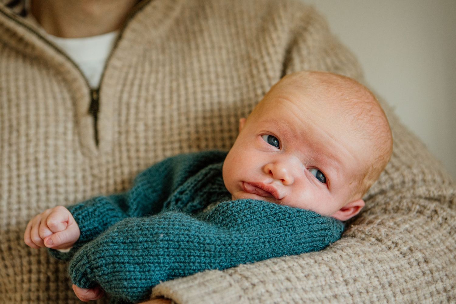 Baby wearing teal knit sweater lying on beige textured fabric.