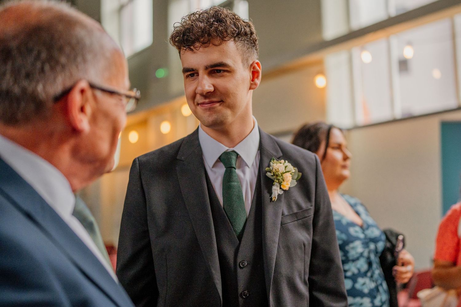 A young person in a dark suit and green tie with boutonniere smiles during a conversation at a formal event.