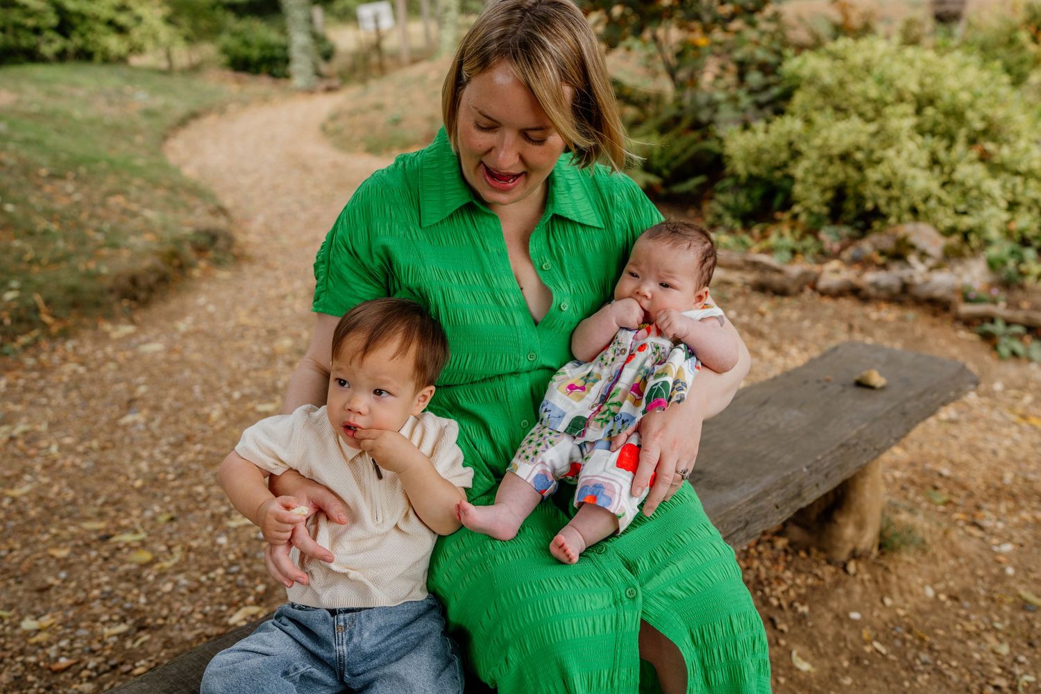 A mother in a green dress sits on a bench outdoors, holding two young babies in a lush garden setting.