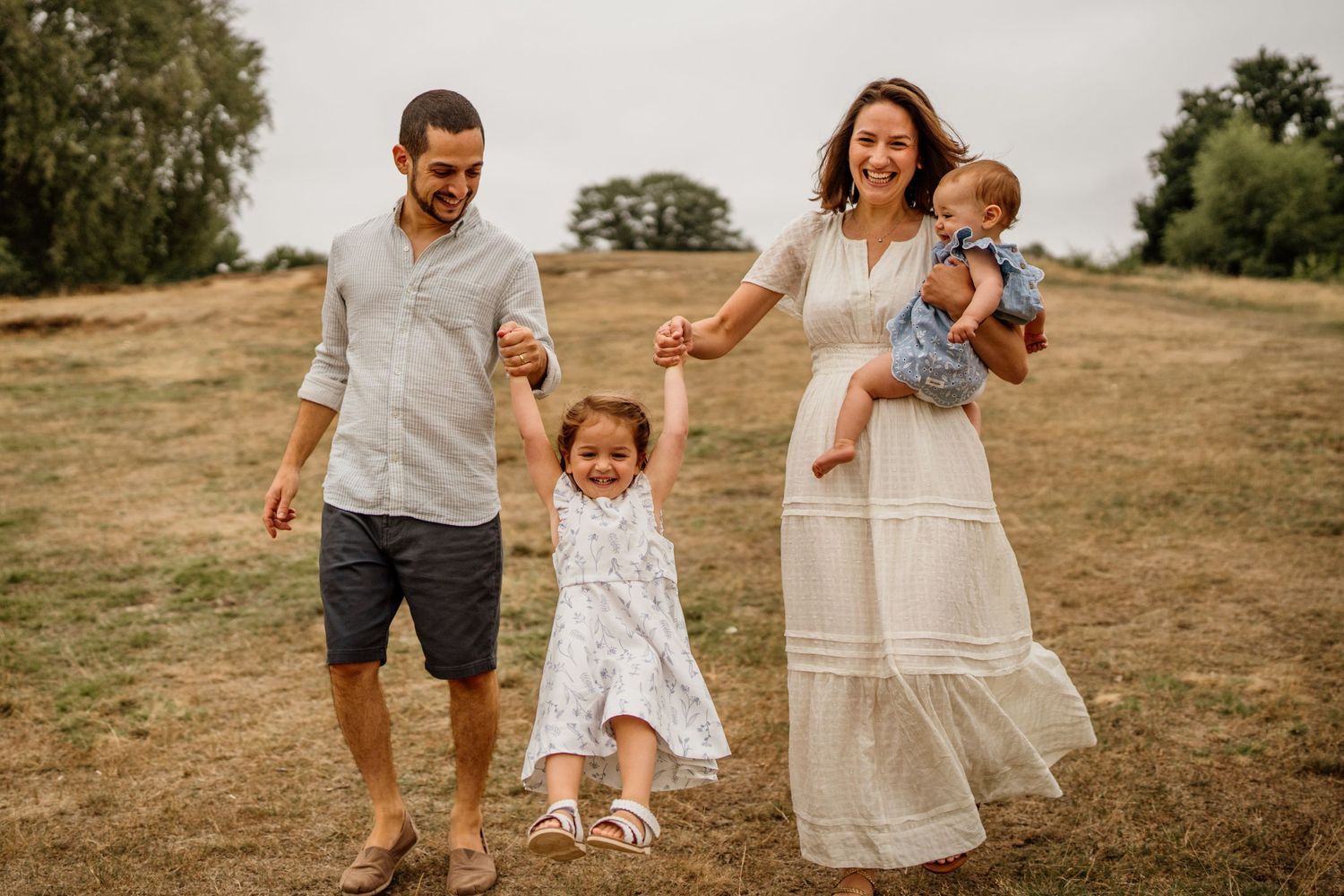 Happy family of four walking in a sunny field, parents swinging toddler girl while mother holds baby boy.