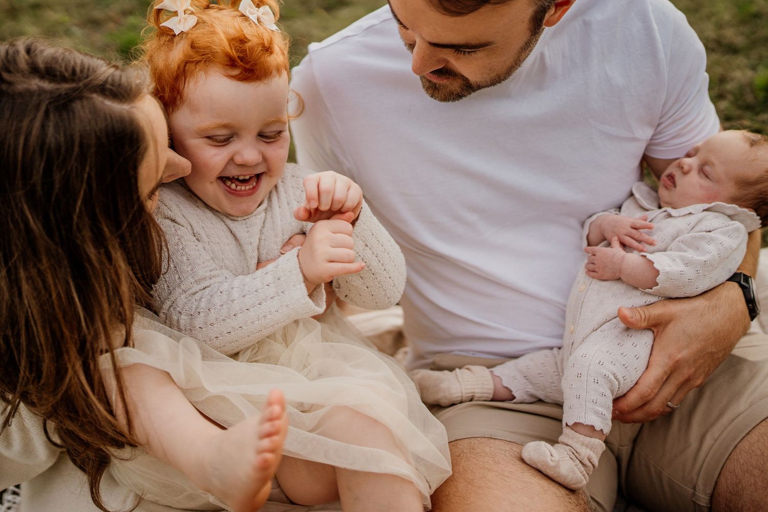 A joyful family sitting outdoors, dad holding a newborn while two young girls laugh and play together.