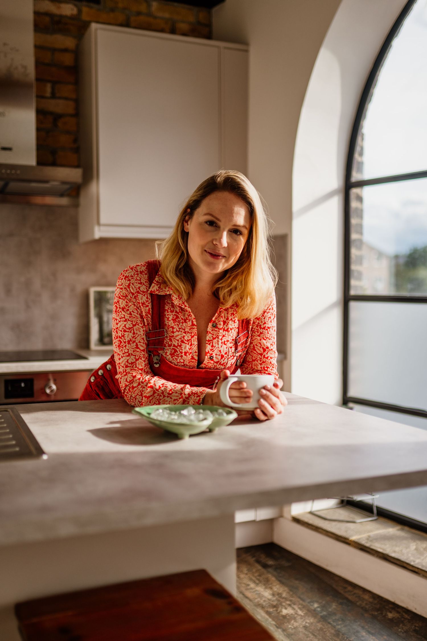 Person in red floral dress leaning on white counter in modern interior with large windows.