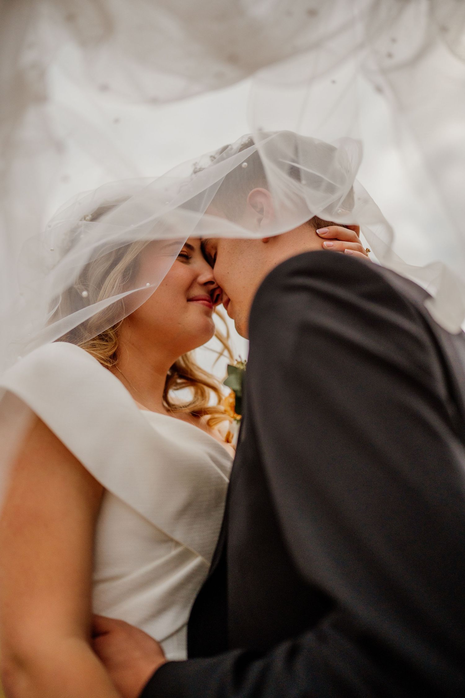 Close-up view of newlyweds embracing, with bride in elegant white off-shoulder dress and flowing veil.