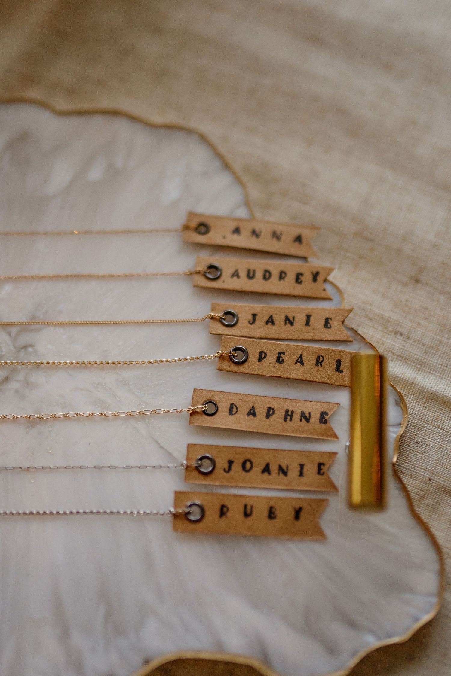 Wooden name tags with handwritten text on delicate chains displayed on light fabric.