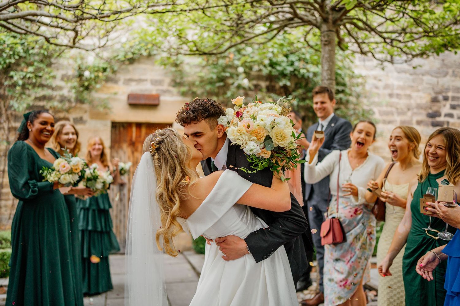 Wedding party celebrates outdoors with bridesmaids in green dresses watching the newlyweds' first kiss.