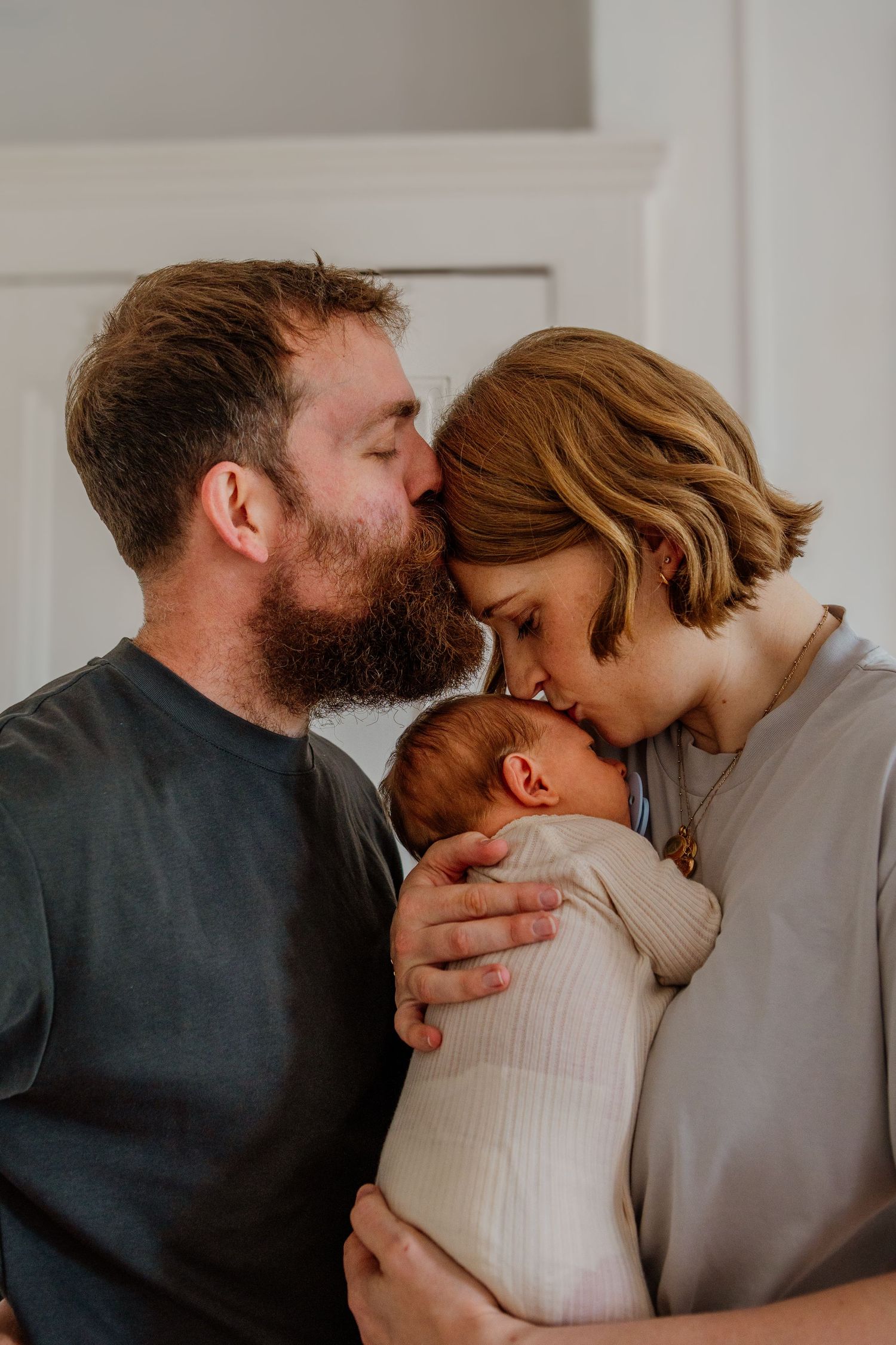 Intimate family moment sharing kisses with baby in white interior setting.