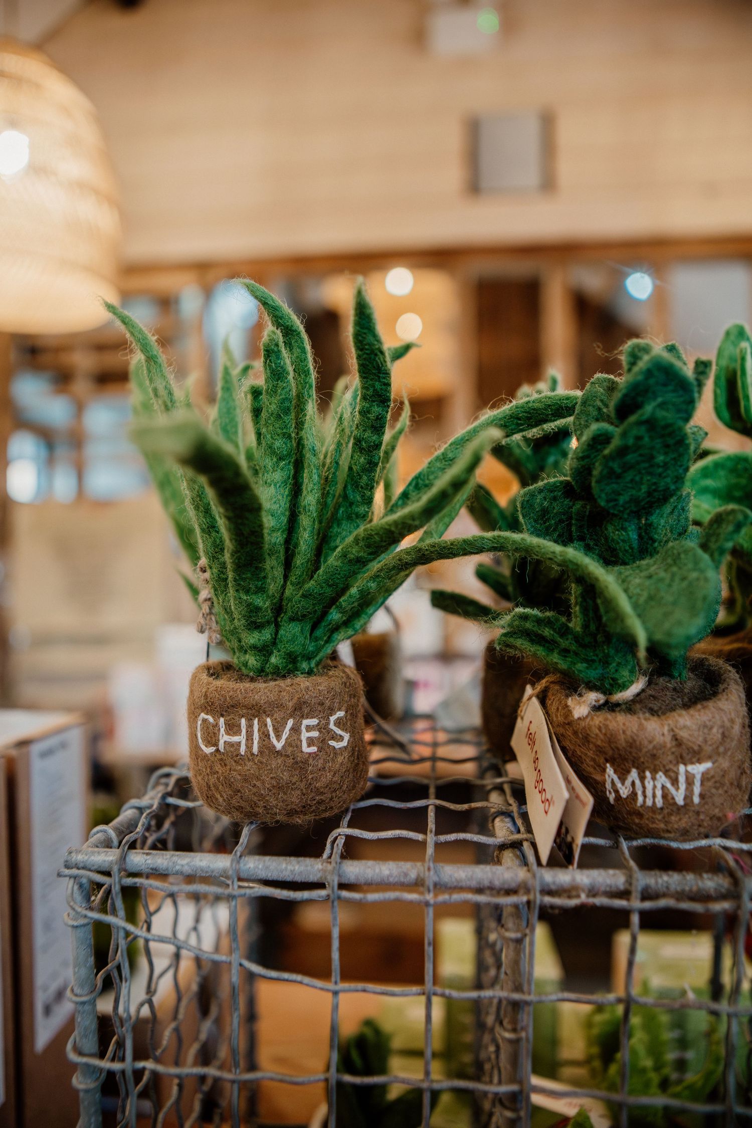 Small potted herbs with chalkboard labels displayed in a rustic wire basket at an indoor market.