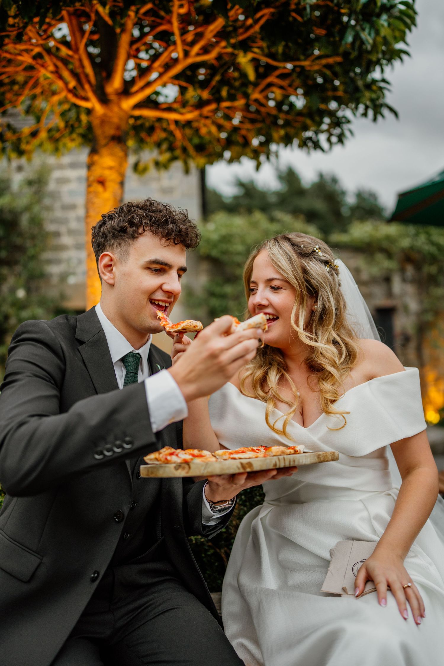 Playful wedding couple shares cake and laughter during outdoor autumn reception celebration.