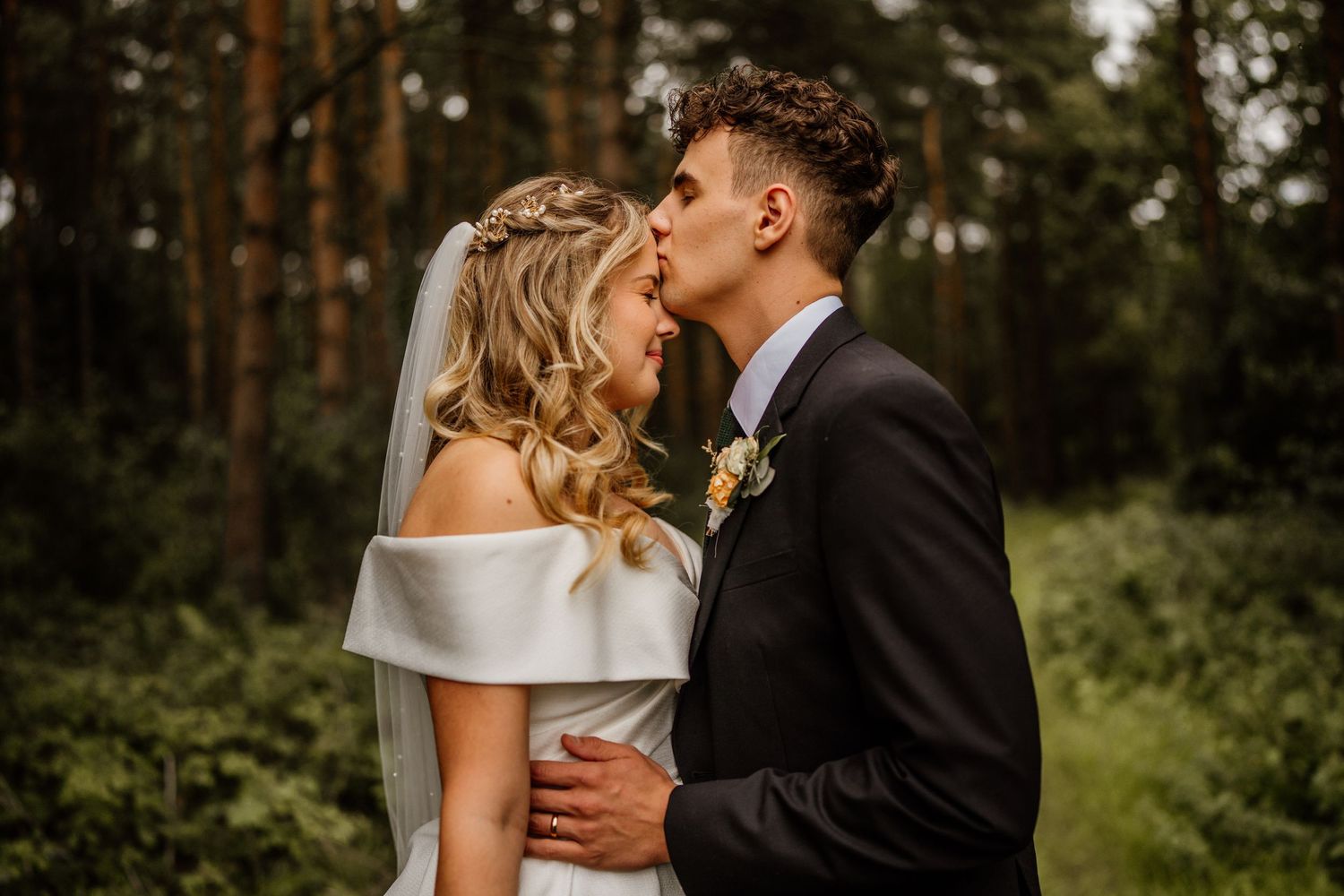 A romantic couple shares a tender kiss in a serene forest setting during their wedding day celebration.