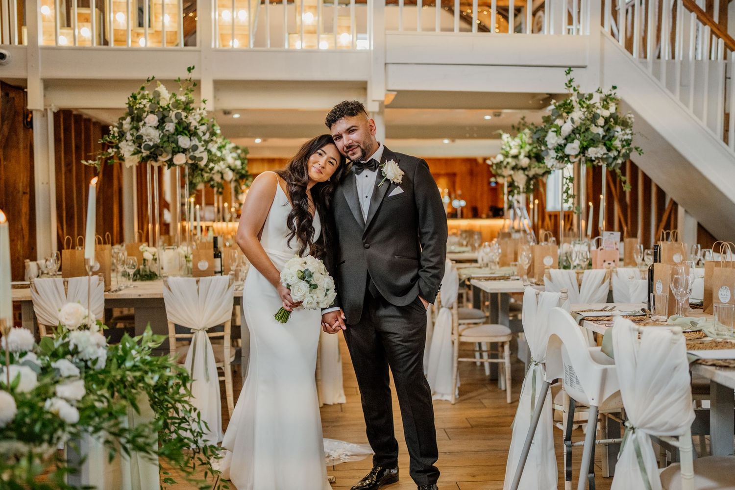 Couple shares a romantic moment in an elegant white wedding venue decorated with floral arrangements and warm lighting.