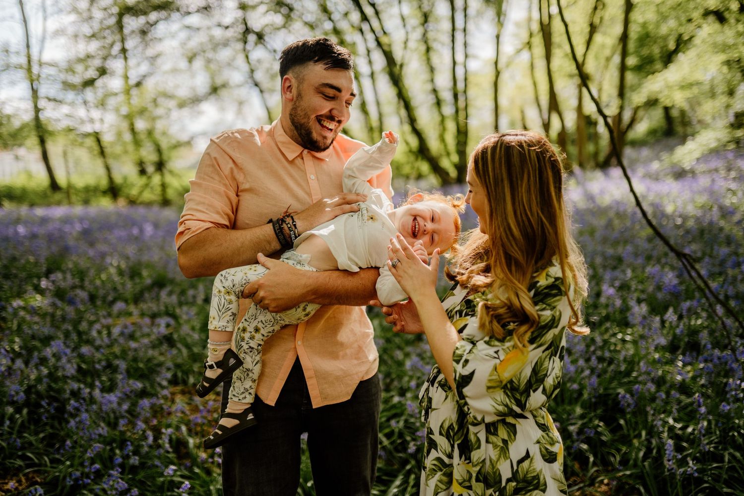 A couple shares a tender moment while holding their baby in a blooming bluebell forest on a sunny day.