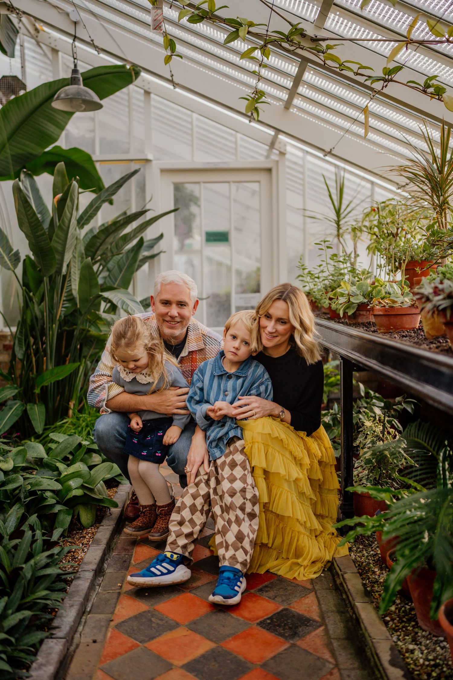 Family gathering in a bright greenhouse with checkered floor and tropical plants.
