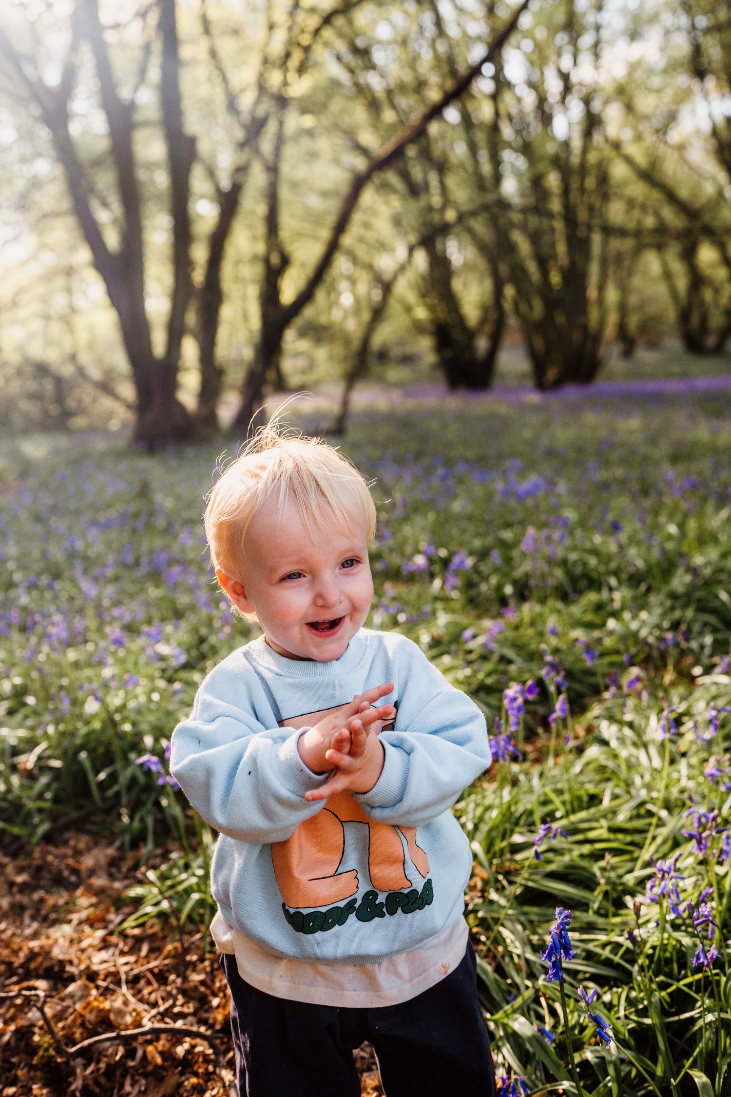 Joyful toddler clapping hands in a bluebell woodland, wearing a light blue sweatshirt on a sunny spring day.