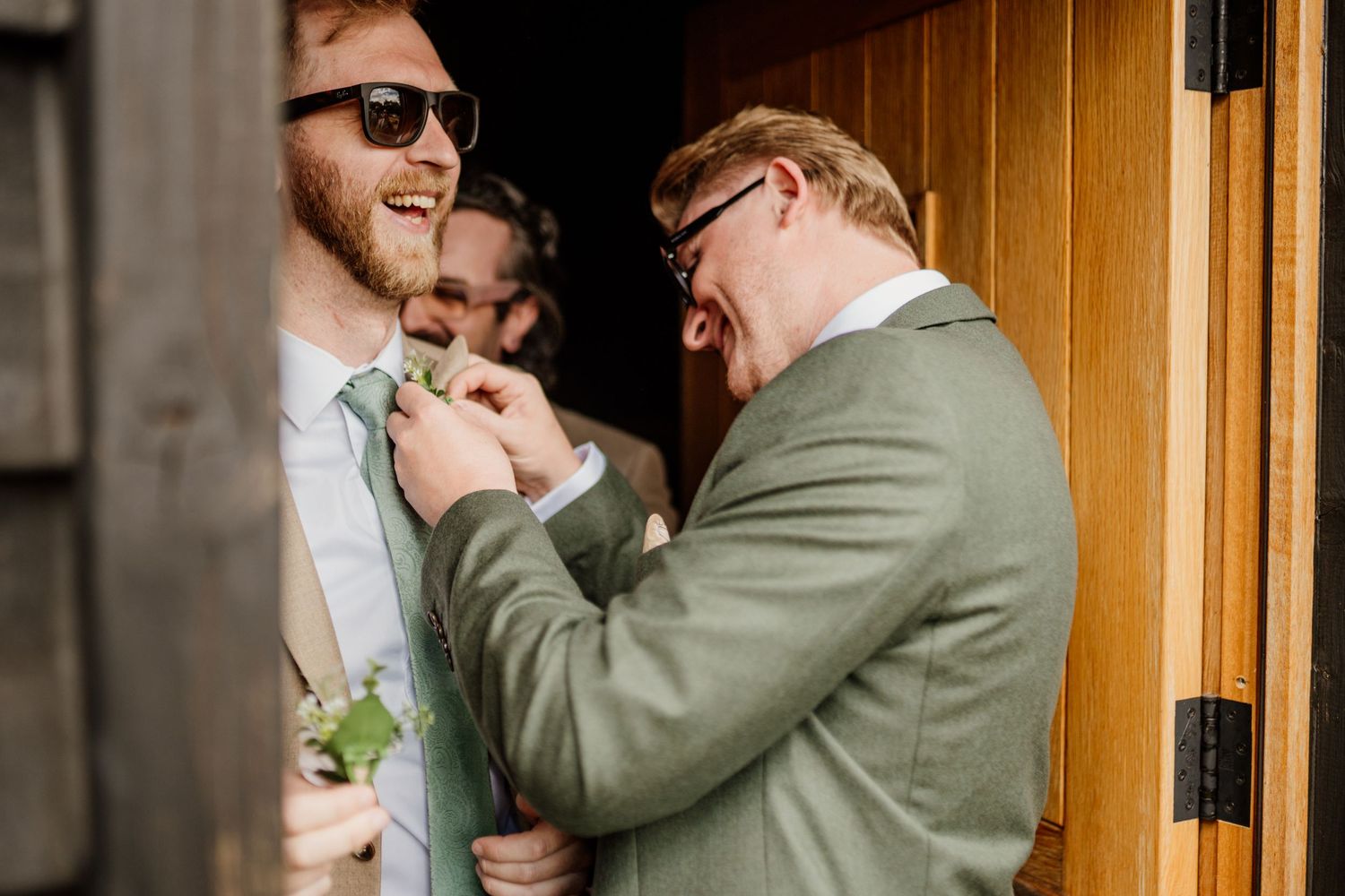 Groomsmen in sunglasses and suits share emotional embraces and laughter in a warmly lit wooden doorway.
